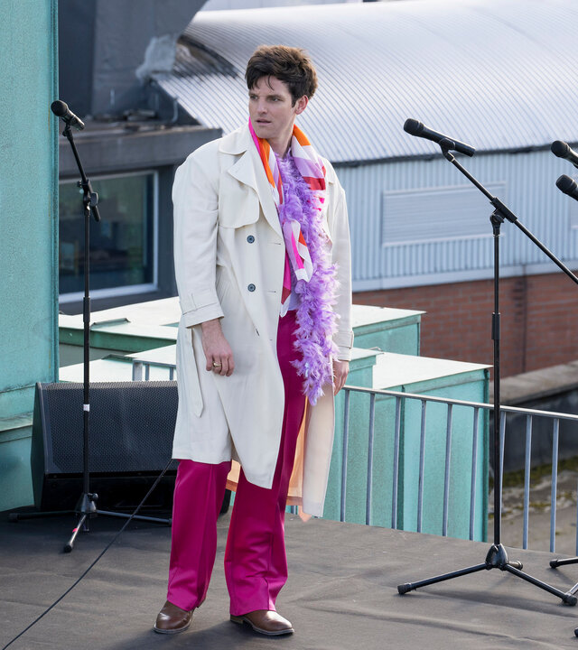 "Harry Wild - Mörderjagd in Dublin - Der seltsame Fall des Jamie Dawson": Jamie Dawson (Adam Weafer) steht auf einem Flachdach. Er ist mit einem weißen Trenchcoat, pinkfarbener Hose und lila Federboa bekleidet. Auf dem Dach neben ihm stehen Mikrofonständer und Boxen.