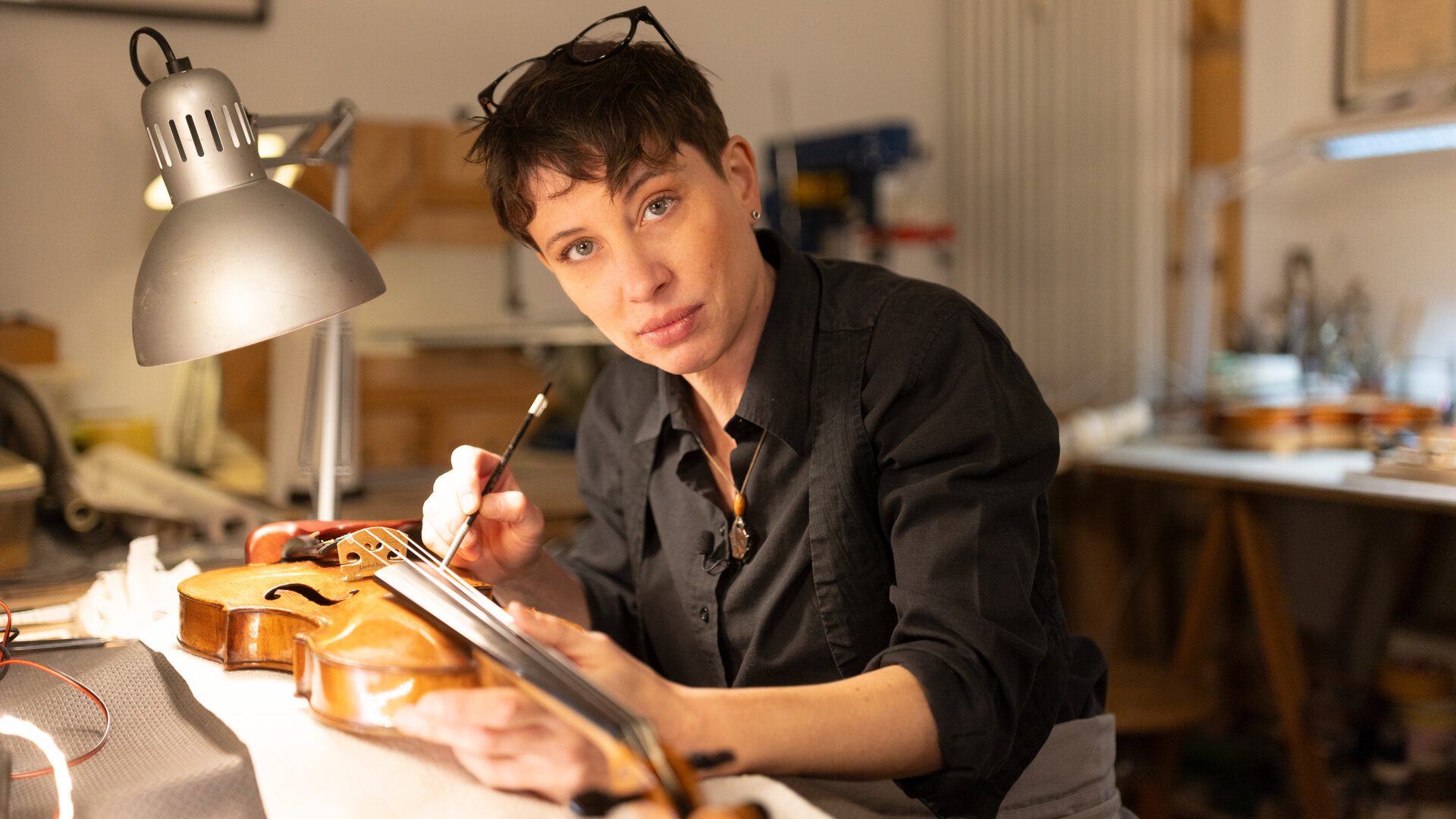 "einfach Mensch - Caroline Tallone: Ich bin mein eigener Chef": Caroline Tallone sitzt an einem Werkstatttisch und arbeitet an einer Geige. Sie hält einen Reinigungspinsel am Griffbrett und schaut dabei in die Kamera. Neben ihr steht eine Lampe, die den Arbeitsplatz beleuchtet.
