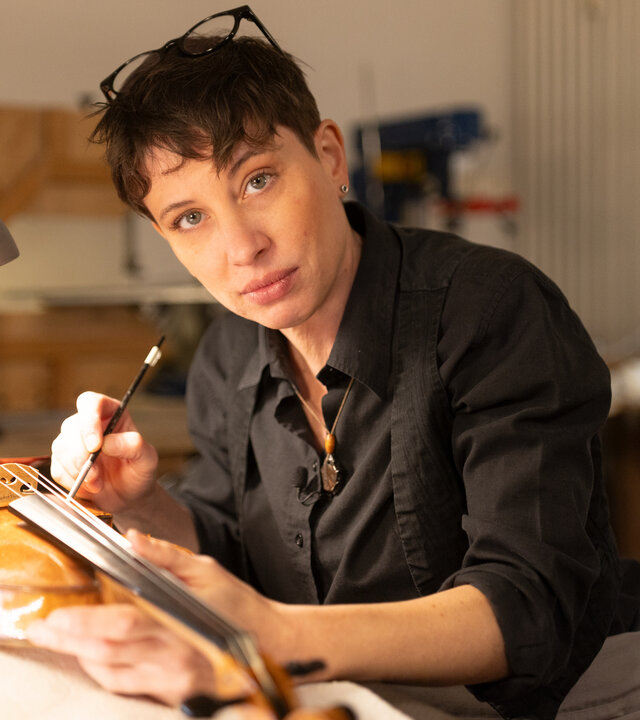 "einfach Mensch - Caroline Tallone: Ich bin mein eigener Chef": Caroline Tallone sitzt an einem Werkstatttisch und arbeitet an einer Geige. Sie hält einen Reinigungspinsel am Griffbrett und schaut dabei in die Kamera. Neben ihr steht eine Lampe, die den Arbeitsplatz beleuchtet.