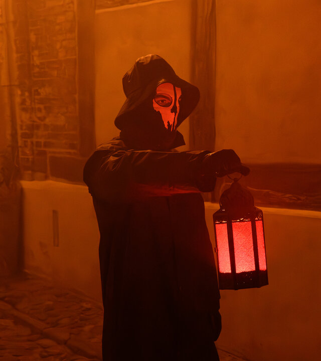 „SOKO Wismar – Der Blüser von Rerik“: In einer Gasse der Wismarer Altstadt steht eine dunkel gekleidete Gestalt, deren Gesicht von einer weißen Geistermaske verdeckt wird. In der erhobenen Hand hält sie eine Laterne, die die nächtliche Gasse in ein rötliches Licht taucht.