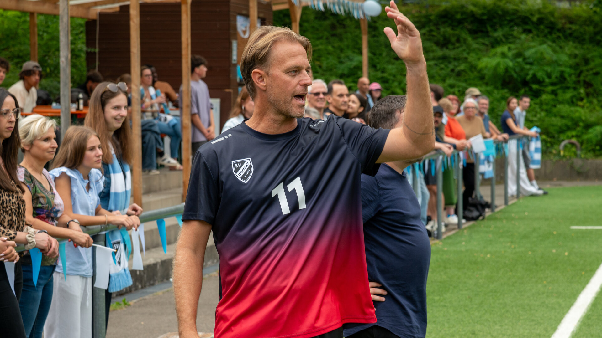 "SOKO Stuttgart - Das Wunder von Zechingen": Chris Aigner (Timo Hildebrand) steht in Trainingskleidung am Rand eines Fußballfeldes und reckt den Arm sich beschwerend in die Höhe. Im Hintergrund befinden sich zahlreiche Zuschauende (Komparsen), die am Spielfeldrand stehen und sitzen.