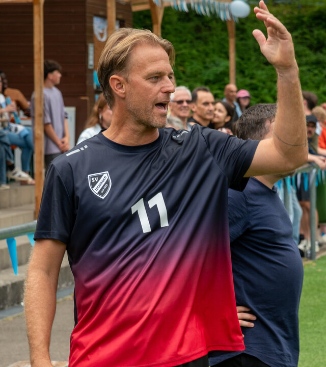"SOKO Stuttgart - Das Wunder von Zechingen": Chris Aigner (Timo Hildebrand) steht in Trainingskleidung am Rand eines Fußballfeldes und reckt den Arm sich beschwerend in die Höhe. Im Hintergrund befinden sich zahlreiche Zuschauende (Komparsen), die am Spielfeldrand stehen und sitzen.