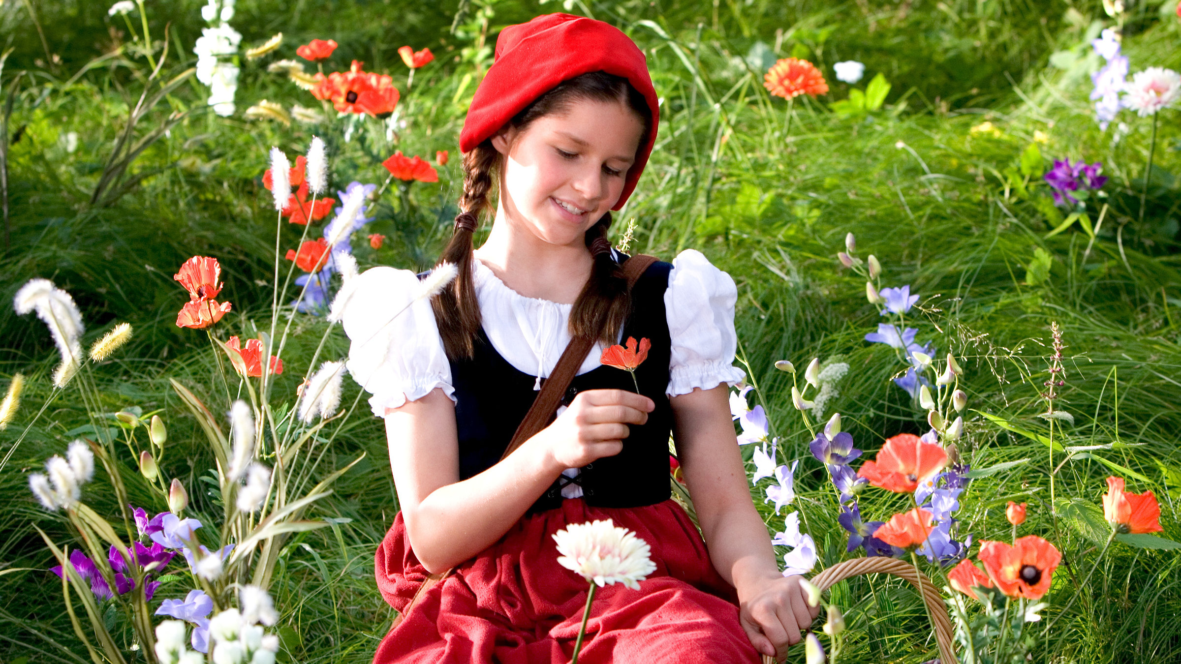 "Rotkäppchen": Rotkäppchen (Kathleen Frontzek) sitzt auf einer Blumenwiese. In der rechten Hand hält sie eine Mohnblume, mit der linken Hand stützt sie sich auf ihrem Körbchen ab. Sie trägt einen roten Rock, eine weiße Bluse und eine kleine schwarze Weste. Auf dem Kopf hat sie eine rote Kappe.