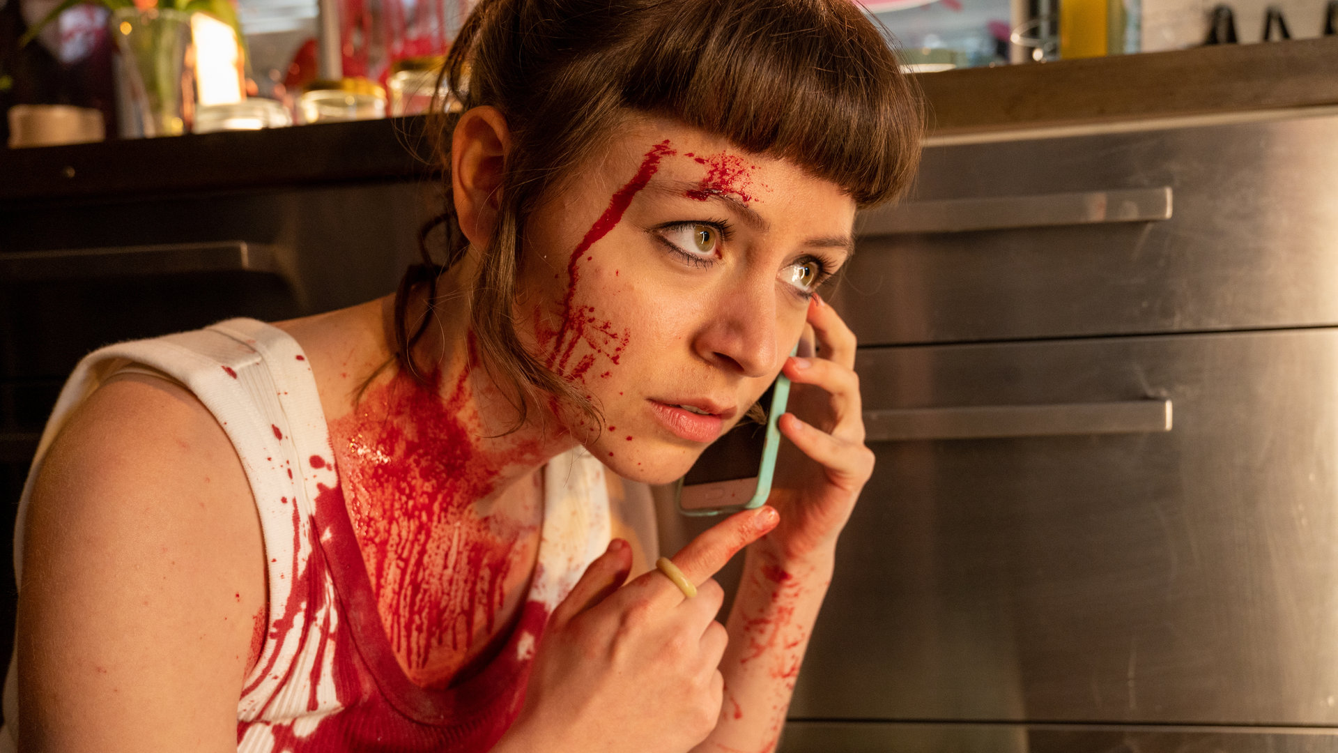 "SOKO Stuttgart - TenderGirl": Paula (Antonia Breidenbach) kauert in ihrem Atelier hinter einer Küchenzeile. Ihr Gesicht und ihr Oberkörper sind blutverschmiert. Sie hält sich ihr Handy ans Ohr. Ihr Gesichtsausdruck ist verzweifelt und ängstlich.