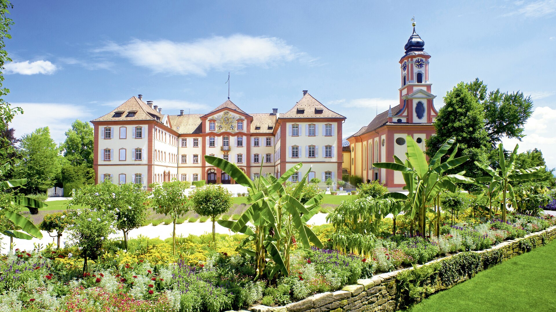 "Evangelischer Gottesdienst - Segel setzen und los": Das Barockschloss auf der Insel Mainau ist von der Sonne beschienen und umgeben von Blumenbeeten, Bäumen und Palmen.