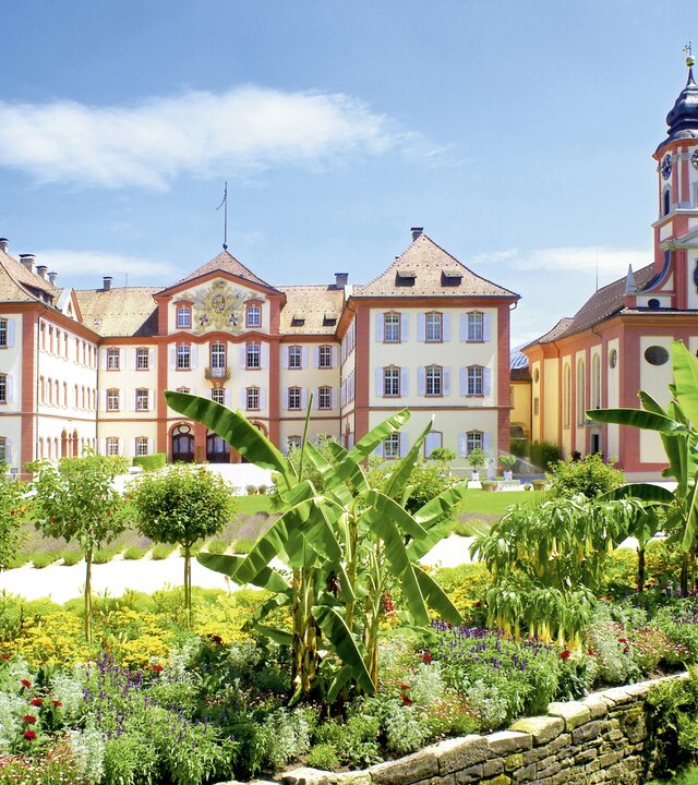 "Evangelischer Gottesdienst - Segel setzen und los": Das Barockschloss auf der Insel Mainau ist von der Sonne beschienen und umgeben von Blumenbeeten, Bäumen und Palmen.