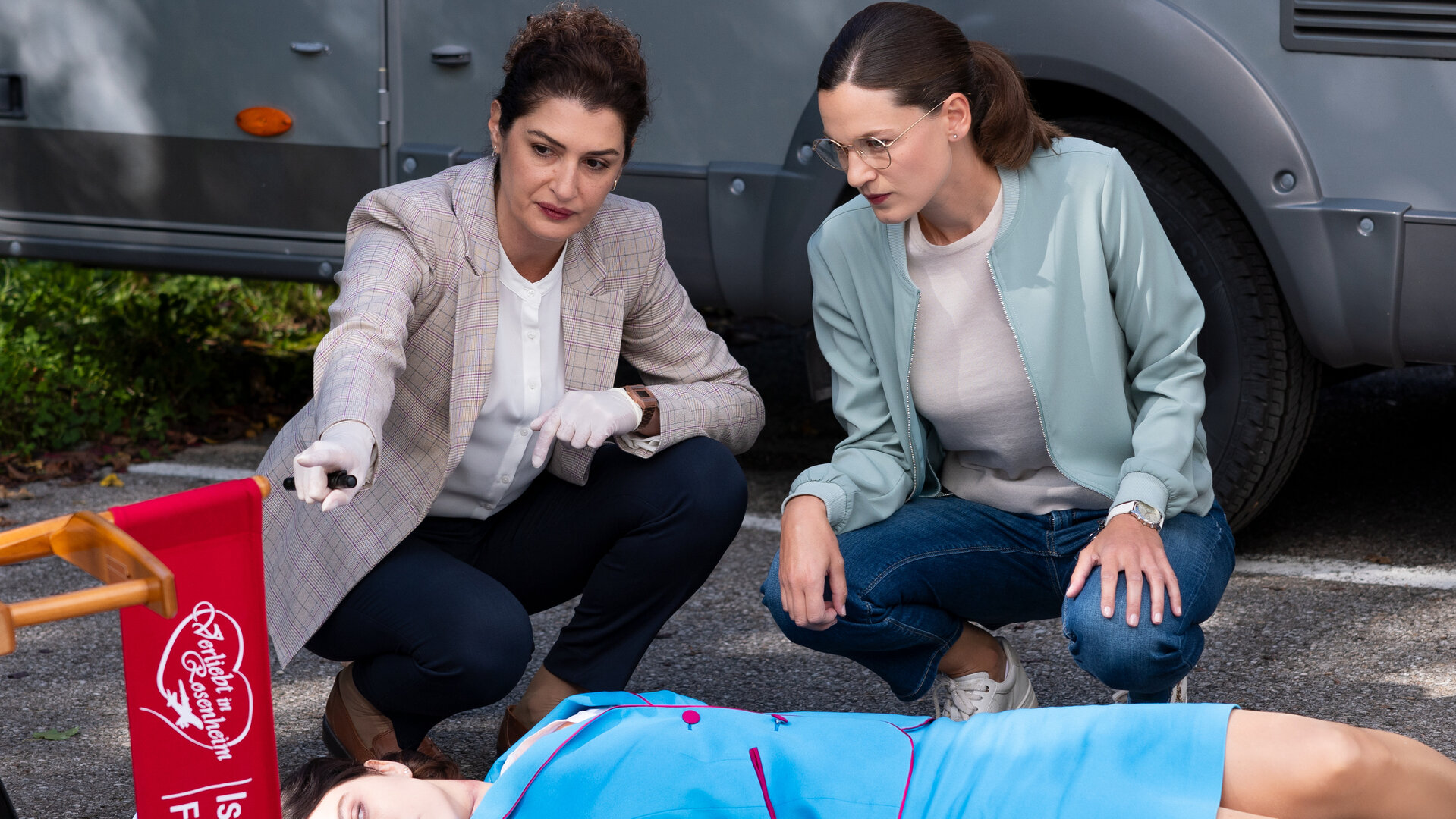 "Die Rosenheim-Cops - Verliebt in Rosenheim": Rechtmedizinerin Dr. Dimos (Anastasia Papadopoulou) und Hauptkommissarin Beck (Michaela Weingartner) hocken vor dem Opfer (Komparsin), das in hellblauem Kostüm rücklings auf dem Asphalt liegt. Daneben ist ein umgekippter roter Regiestuhl zu sehen, der die weiße Aufschrift "Verliebt in Rosenheim - Isabell Feichtel" trägt.