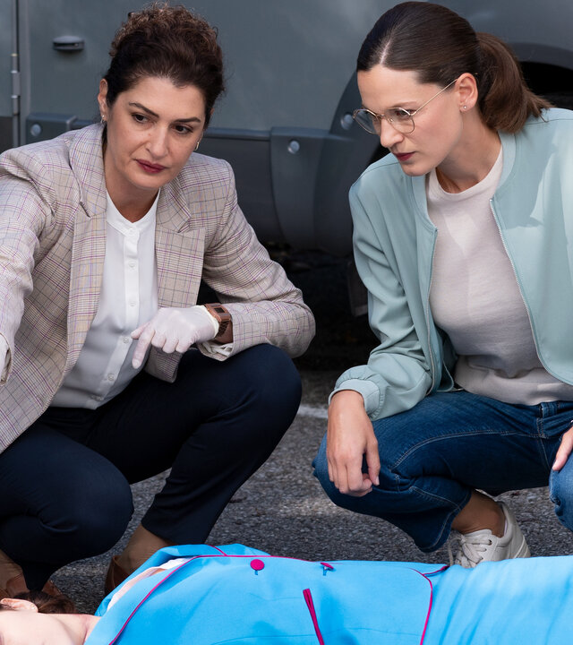 "Die Rosenheim-Cops - Verliebt in Rosenheim": Rechtmedizinerin Dr. Dimos (Anastasia Papadopoulou) und Hauptkommissarin Beck (Michaela Weingartner) hocken vor dem Opfer (Komparsin), das in hellblauem Kostüm rücklings auf dem Asphalt liegt. Daneben ist ein umgekippter roter Regiestuhl zu sehen, der die weiße Aufschrift "Verliebt in Rosenheim - Isabell Feichtel" trägt.