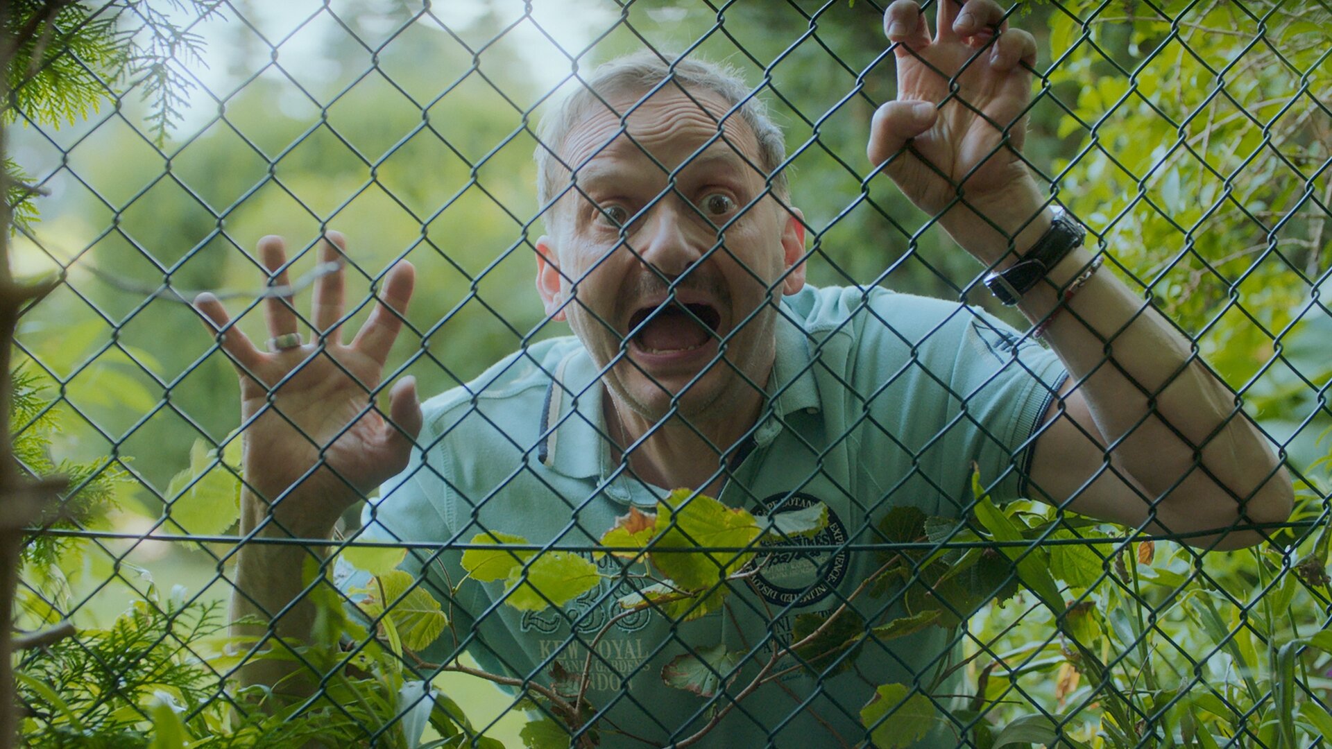 "Doppelhaushälfte - Ortsversteher": Andi (Milan Peschel) steht mit theatralischem Blick und weit aufgerissenen Augen sowie offenem Mund hinter einem Drahtzaun, den er mit seinen Händen festhält.