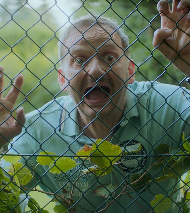"Doppelhaushälfte - Ortsversteher": Andi (Milan Peschel) steht mit theatralischem Blick und weit aufgerissenen Augen sowie offenem Mund hinter einem Drahtzaun, den er mit seinen Händen festhält.