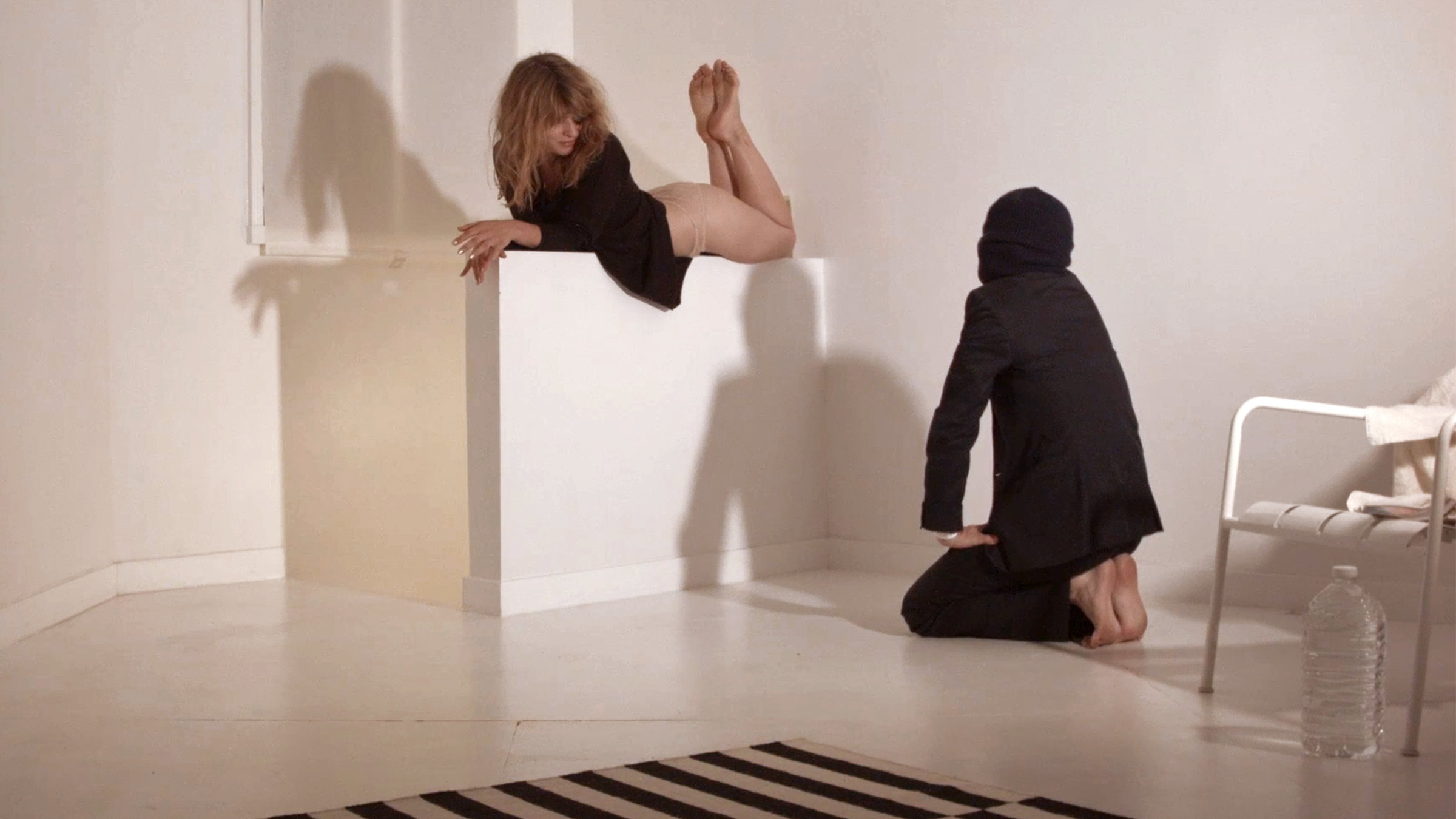 Ein modernes Atelier: eine junge Frau räkelt sich in lasziver Pose auf einer Mauer, während zu ihren Füßen ein vermummter Mann im Anzug kauert.