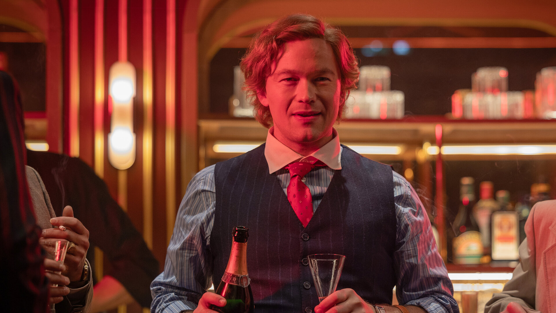 "Stenbeck - Zwischen zwei Welten": Jan Stenbeck (Jakob Oftebro) steht mit Sektflasche und Sektglas in Händen vor einer rötlich beleuchteten Bar.