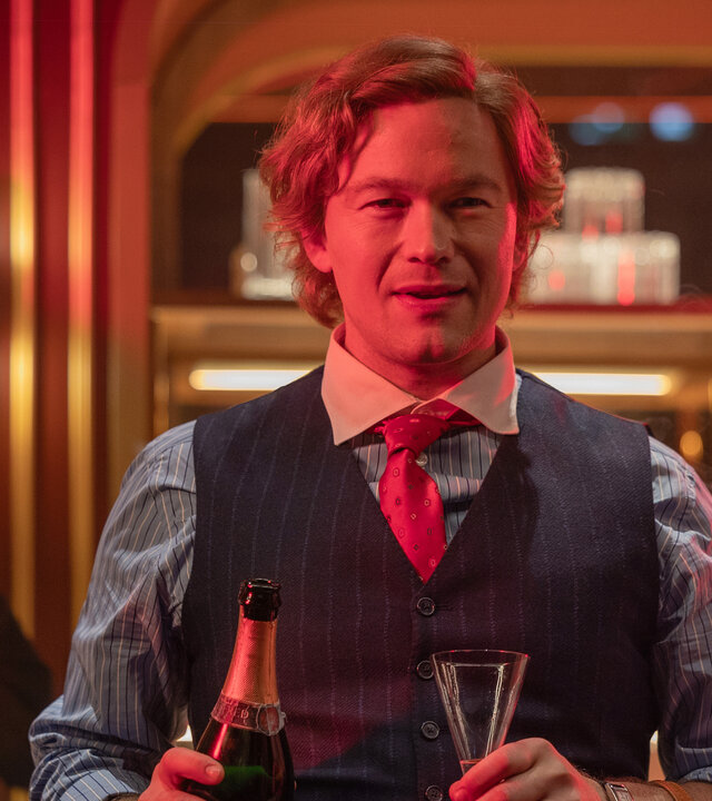"Stenbeck - Zwischen zwei Welten": Jan Stenbeck (Jakob Oftebro) steht mit Sektflasche und Sektglas in Händen vor einer rötlich beleuchteten Bar.