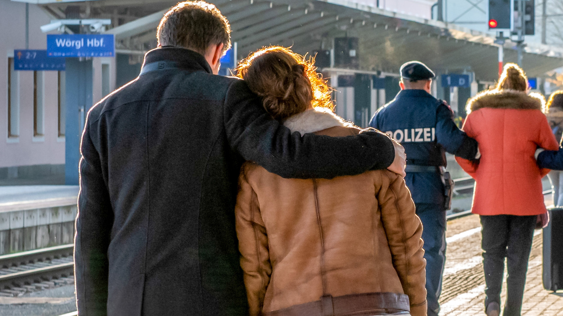 "SOKO Kitzbühel - Abschied": An einem Bahnsteig läuft Lukas (Jakob Seeböck) mit Nina (Julia Cencig) entlang. Er legt seinen Arm um ihre Schulter. Vor ihnen wird eine Frau von der Polizei begleitet. Alle Personen sind von hinten zu sehen.