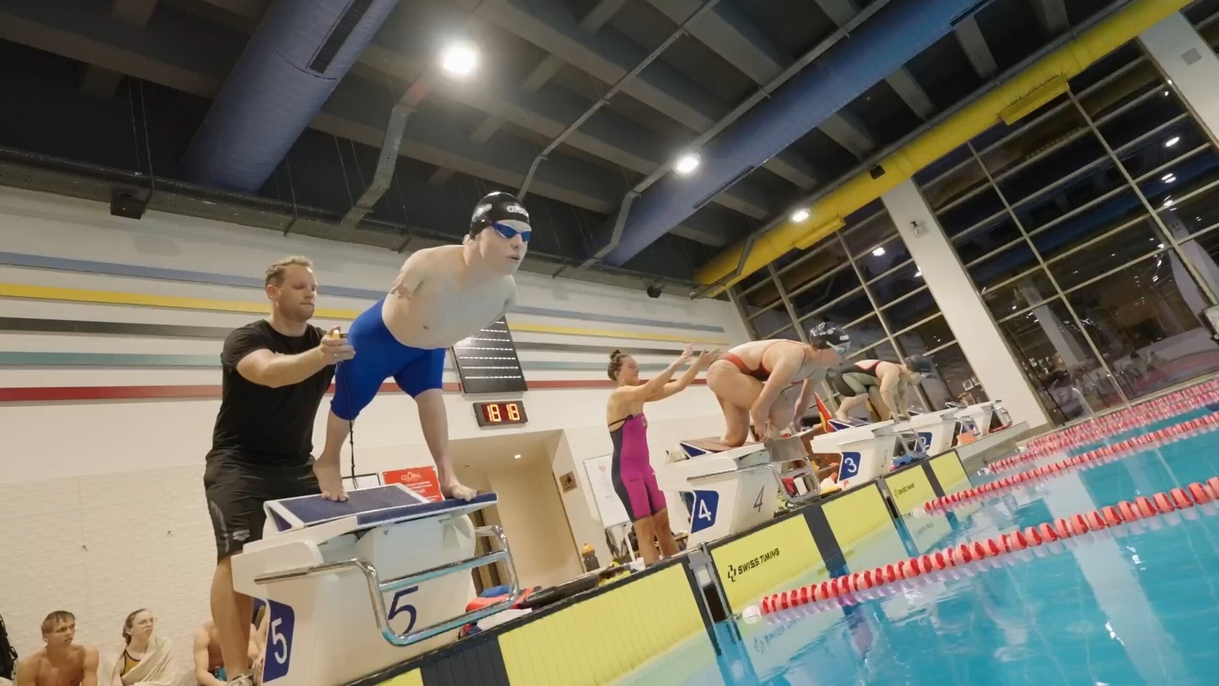 "einfach Mensch - Josia Topf: Schwimmer gegen alle Widerstände (4)": Im Schimmbad: Josia Topf startet bei einem Schwimmwettkampf mit einem Sprung vom Startblock ins Wasser. Im Hintergrund starten zwei weitere Schimmer.