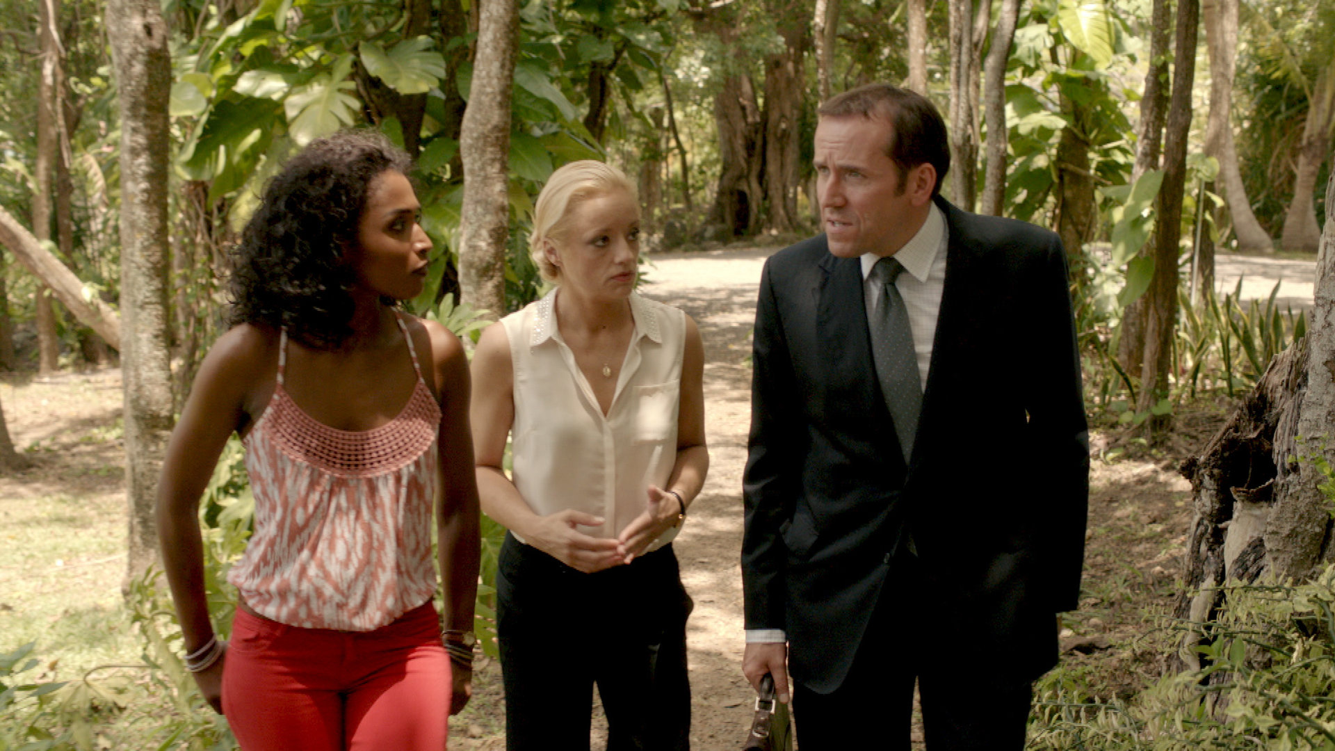 "Death in Paradise - Ein (fast) perfekter Plan": Camille Bordey (Sara Martins), Vicky Woodward (Lucy Davis) schauen gespannt DI Richard Poole (Ben Miller) an, der mit ihnen redet. Alle drei laufen nebeneinander im Freien einen Weg entlang.