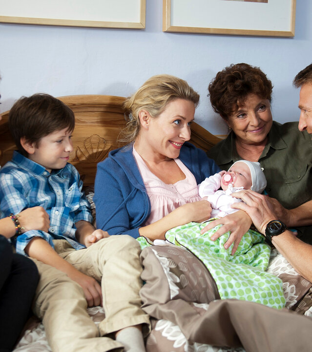 "Der Bergdoktor: Keine Zukunft": Im Schlafzimmer: Glücklich lächelnd blicken sich Susanne Dreiseitl (Nathalie O'Hara) und Hans Gruber (Heiko Ruprecht) an. Susanne sitzt im Bett und hält ein Baby ihm Arm. Rechts von ihr sitzen Jonas (Fabian Elias Huber) und Lilli (Ronja Forcher), links von ihr Lisbeth (Monika Baumgartner). Alle lächeln freudig.