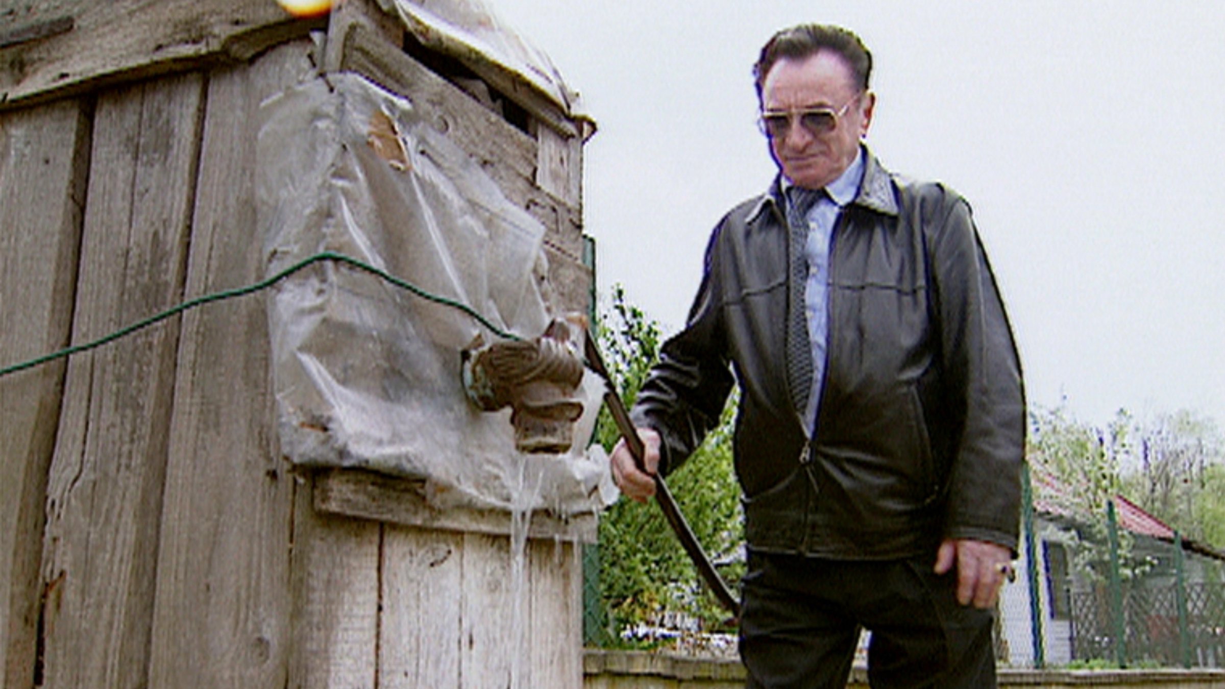 "Die Verwandlung des guten Nachbarn": Ein älterer Mann mit schwarzer Lederjacke, Krawatte und Sonnenbrille steht an der Pumpe eines Brunnens und bewegt den Hebel.