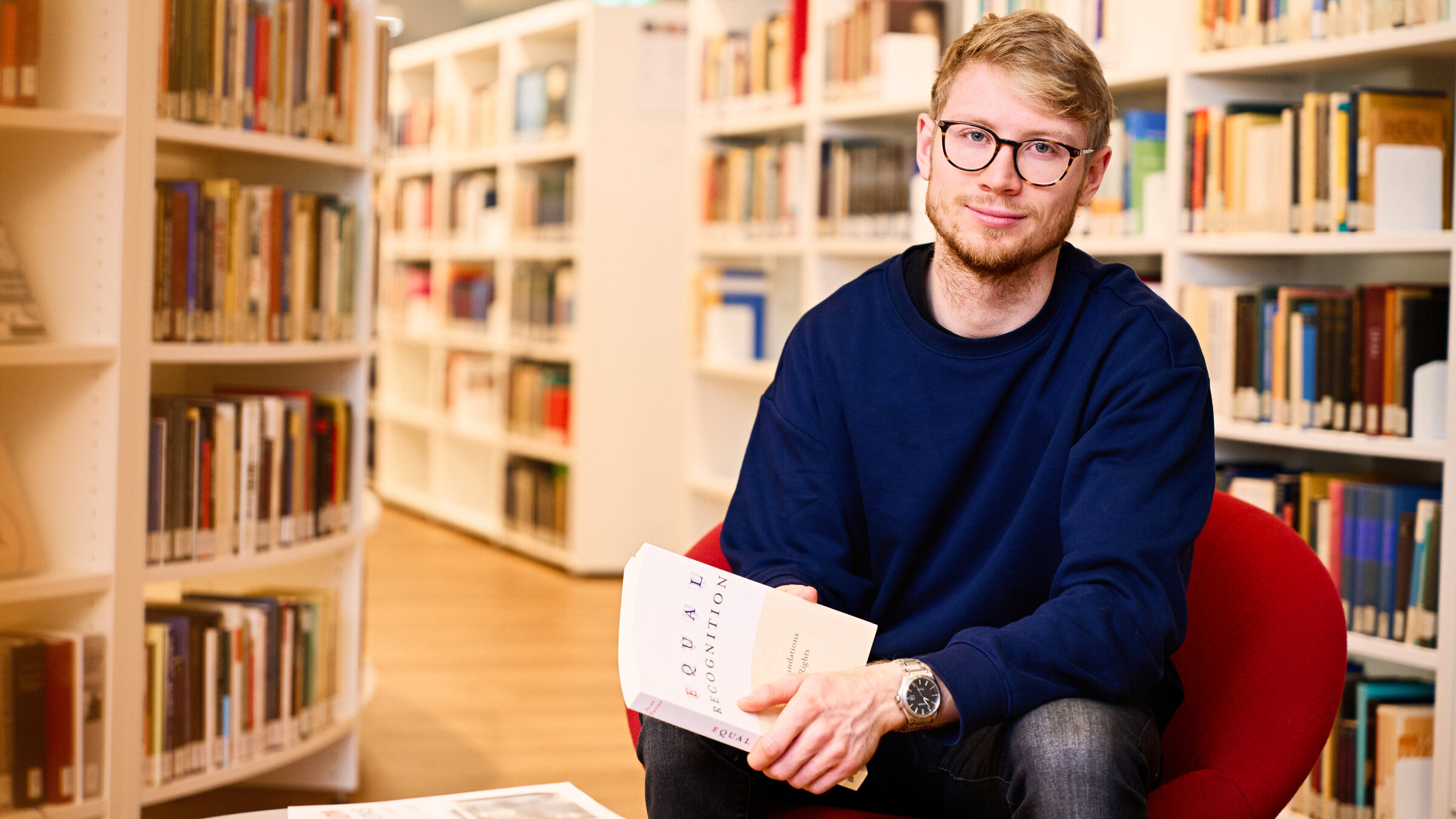 "einfach Mensch - Moritz Raykowski: Ich forsche für mehr Menschenrechte!": In einer Bibliothek: Moritz sitzt auf einem roten Sessel. Er hält ein Buch in der Hand und schaut lächelnd in die Kamera.
