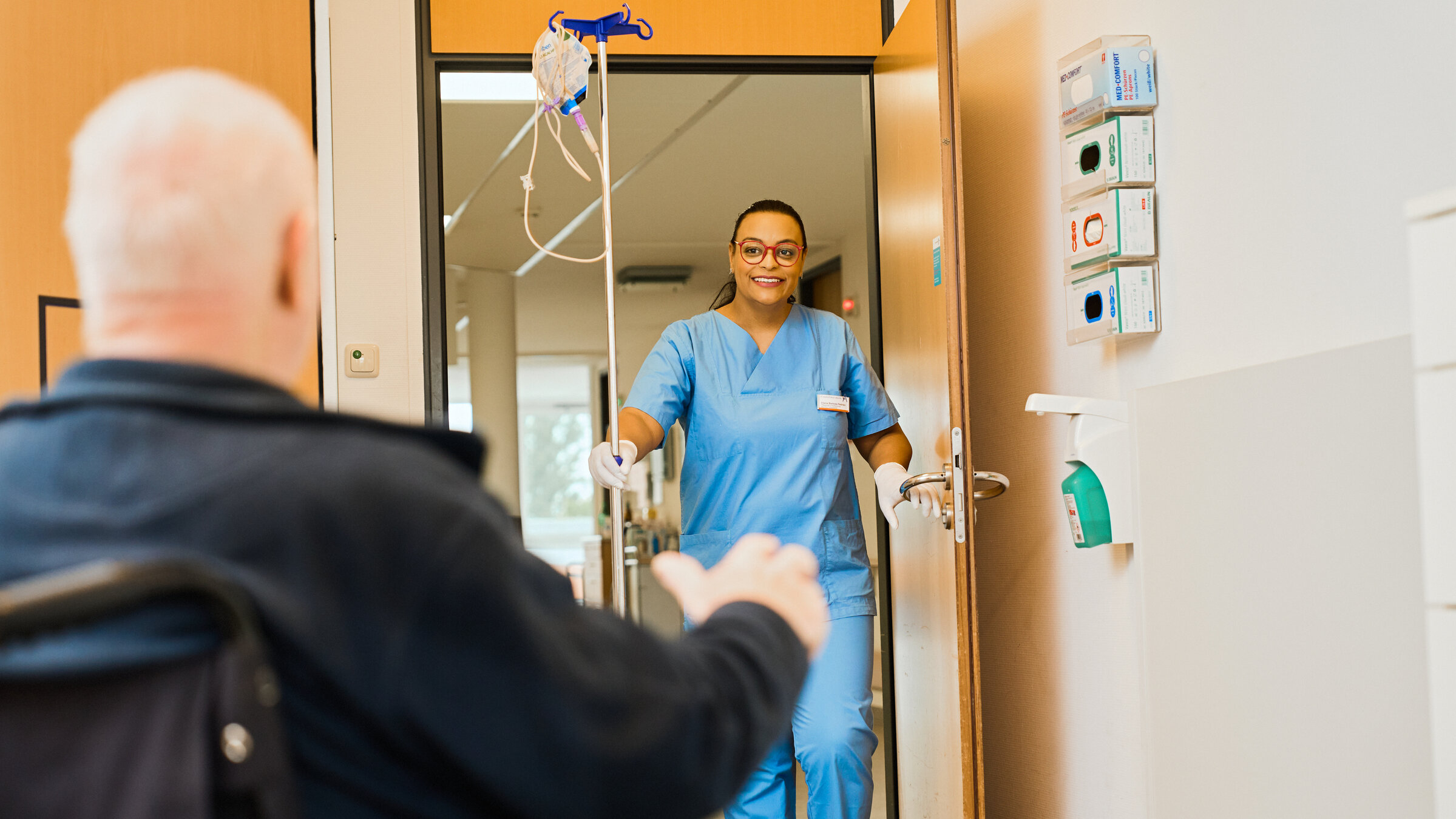 "37°: Arbeitskräfte weltweit gesucht!": Im Krankenhaus: Elaine, die Pflegekraft steht lächelnd in der offenen Tür. Ein älterer Mann im sitzt, mit dem Rücken zur Kamera, im Rollstuhl in einem Zimmer.