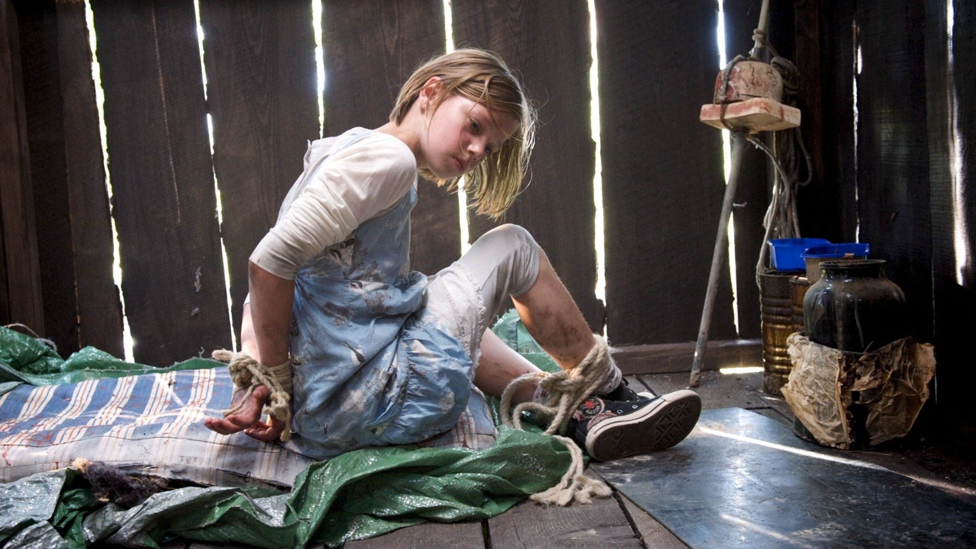 "SOKO Wismar - Flaschenpost": Die kleine Klara Husemann (Hanna Höppner) sitzt in einer Holzhütte auf einer grünen Plane und einer blau-weißen Decke. Ihre Handgelenke sind auf dem Rücken gefesselt, und ihre Füße sind aneinander gebunden. Mit einer großen Stange wird die Hütte geschlossen gehalten.