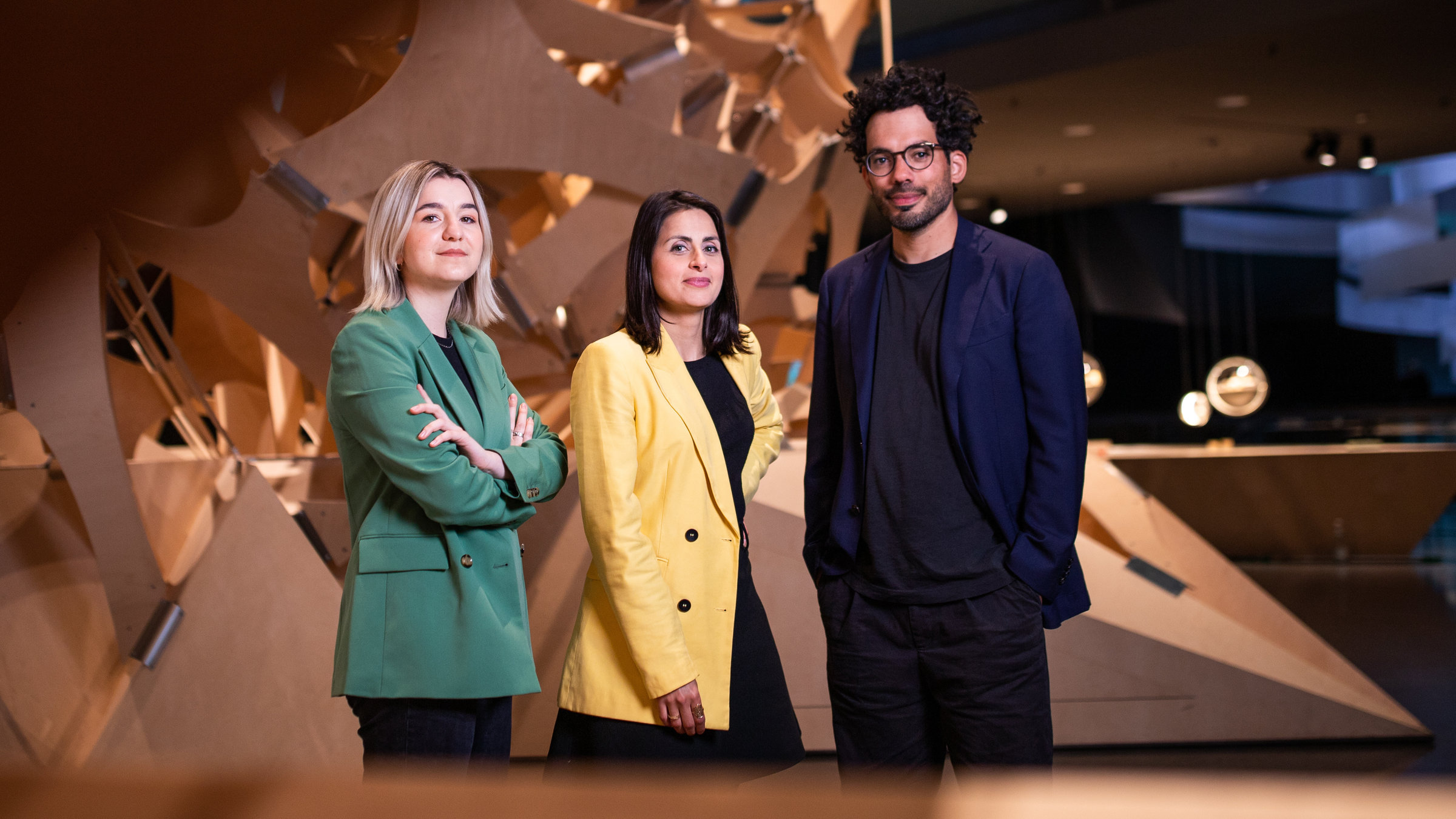 "Futur Wir – Das neue deutsche Selbstverständnis (1/3) - Wer wir sein wollen": Die drei Moderatoren stehen im Berliner Futurium und blicken in die Kamera. Eingerahmt sind sie von einer großen Holzskulptur.