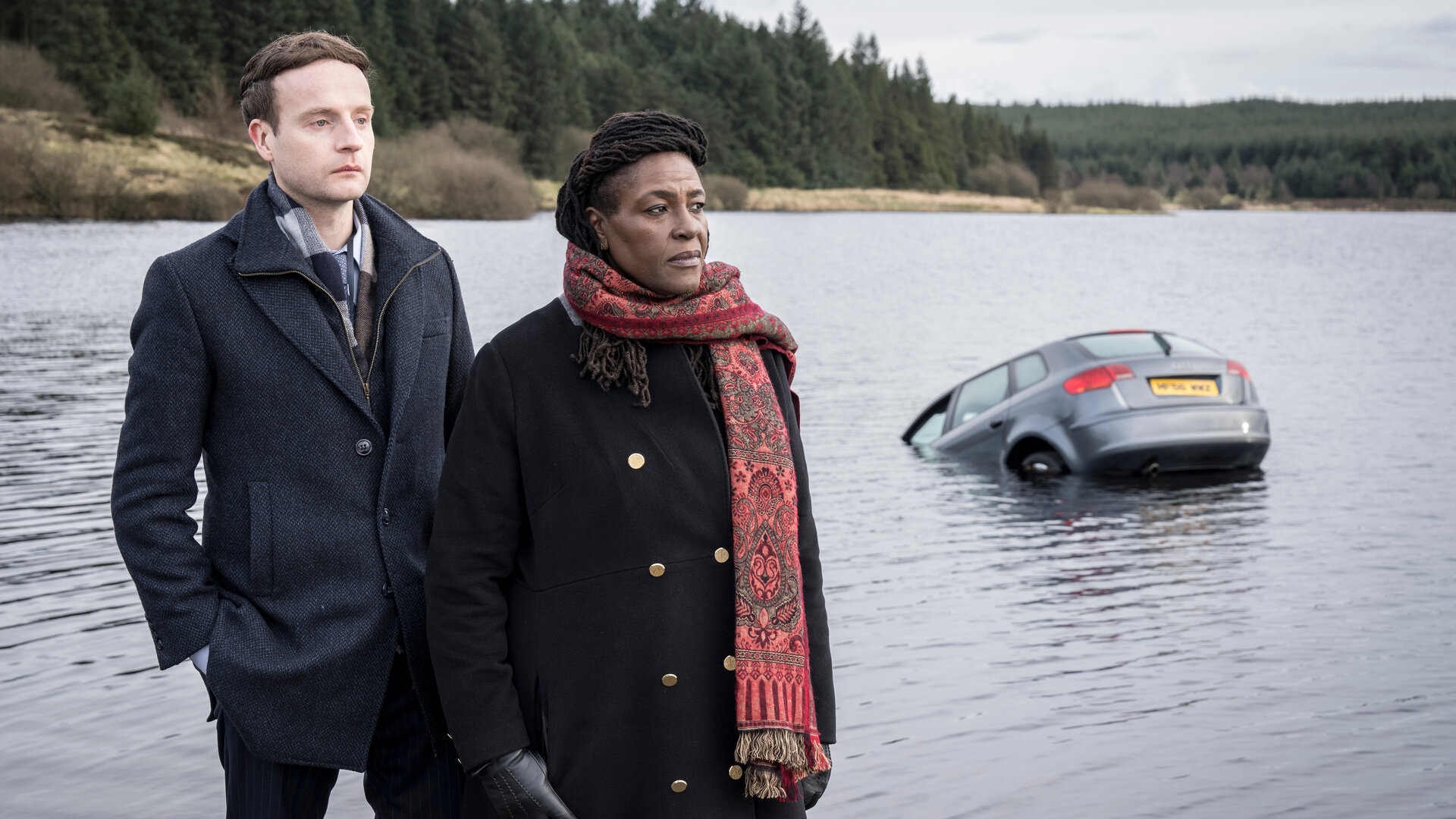 "Ellis - Blut und Wasser": DS Harper (Andrew Gower) und DCI Ellis (Sharon D Clarke), eine Frau mit jamaikanischen Wurzeln, stehen am Ufer eines Gewässers, aus dem die hintere Hälfte eines Autos herausragt. Beide schauen zur Seite. Das gegenüberliegende Ufer ist bewaldet.
