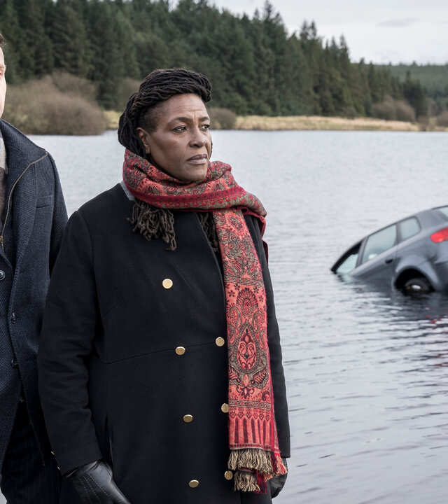 "Ellis - Blut und Wasser": DS Harper (Andrew Gower) und DCI Ellis (Sharon D Clarke), eine Frau mit jamaikanischen Wurzeln, stehen am Ufer eines Gewässers, aus dem die hintere Hälfte eines Autos herausragt. Beide schauen zur Seite. Das gegenüberliegende Ufer ist bewaldet.