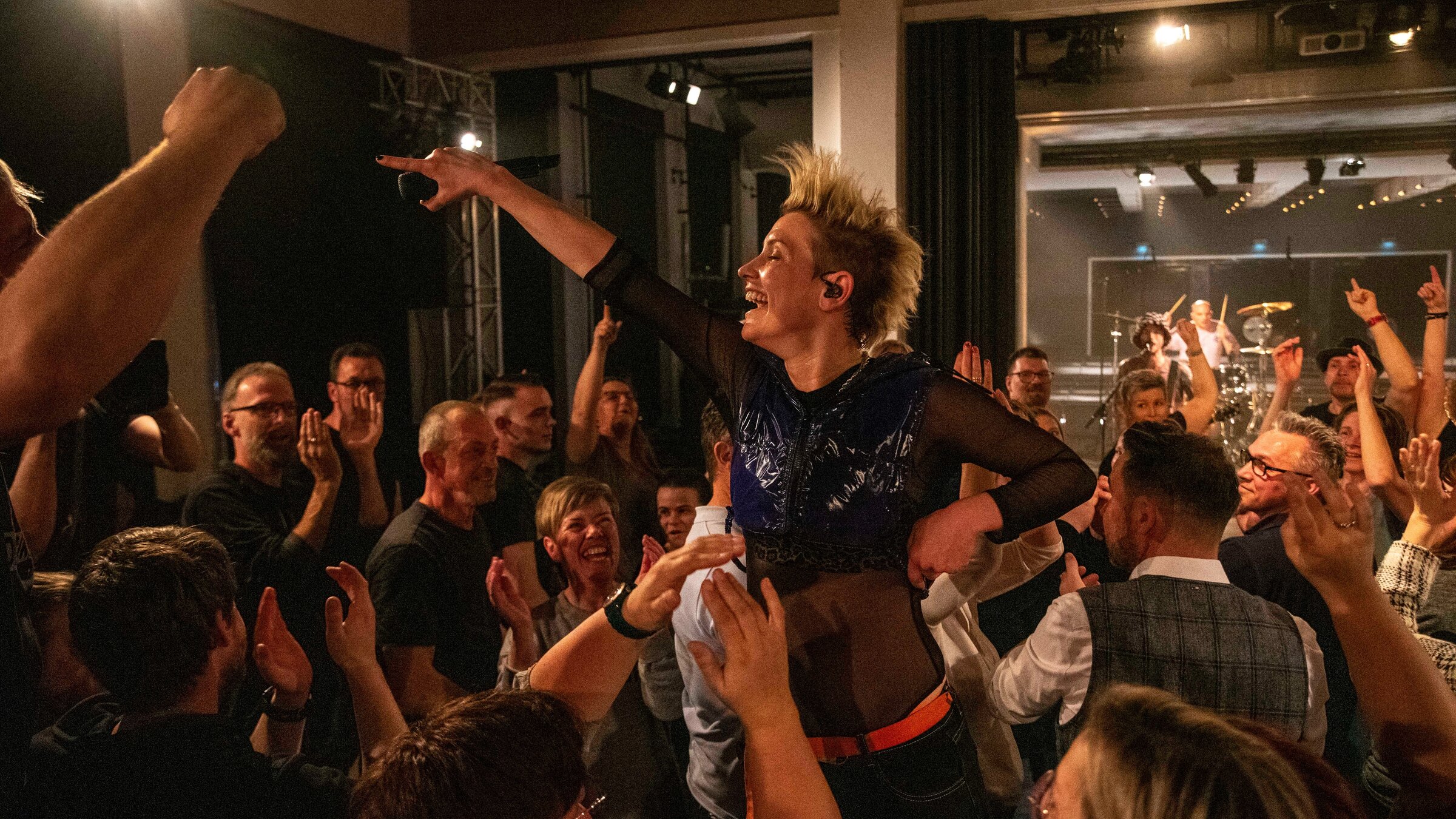 "Pop Around@Bauhaus: Deine Cousine": Die Sängerin steht inmitten einer Gruppe von Konzertbesuchern, die teils klatschen oder die Arme in die Luft strecken. Die Sängerin hat den linken Arm nach oben gestreckt. Sie ist gekleidet mit einem schwarzen, paillettenbesetzten Oberteil. Im Hintergrund ist die Bühne mit einem Schlagzeuger zu erkennen.