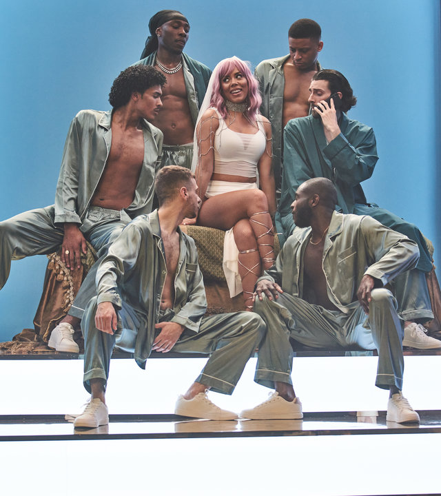 "Mood: Get That Schmoneyyy": Sasha (Nicôle Lecky) sitzt auf einer kleinen Showtreppe. Sie ist weiß gekleidet und trägt eine rosa Perücke. Sechs Männer umringen sie und blicken in ihre Richtung.