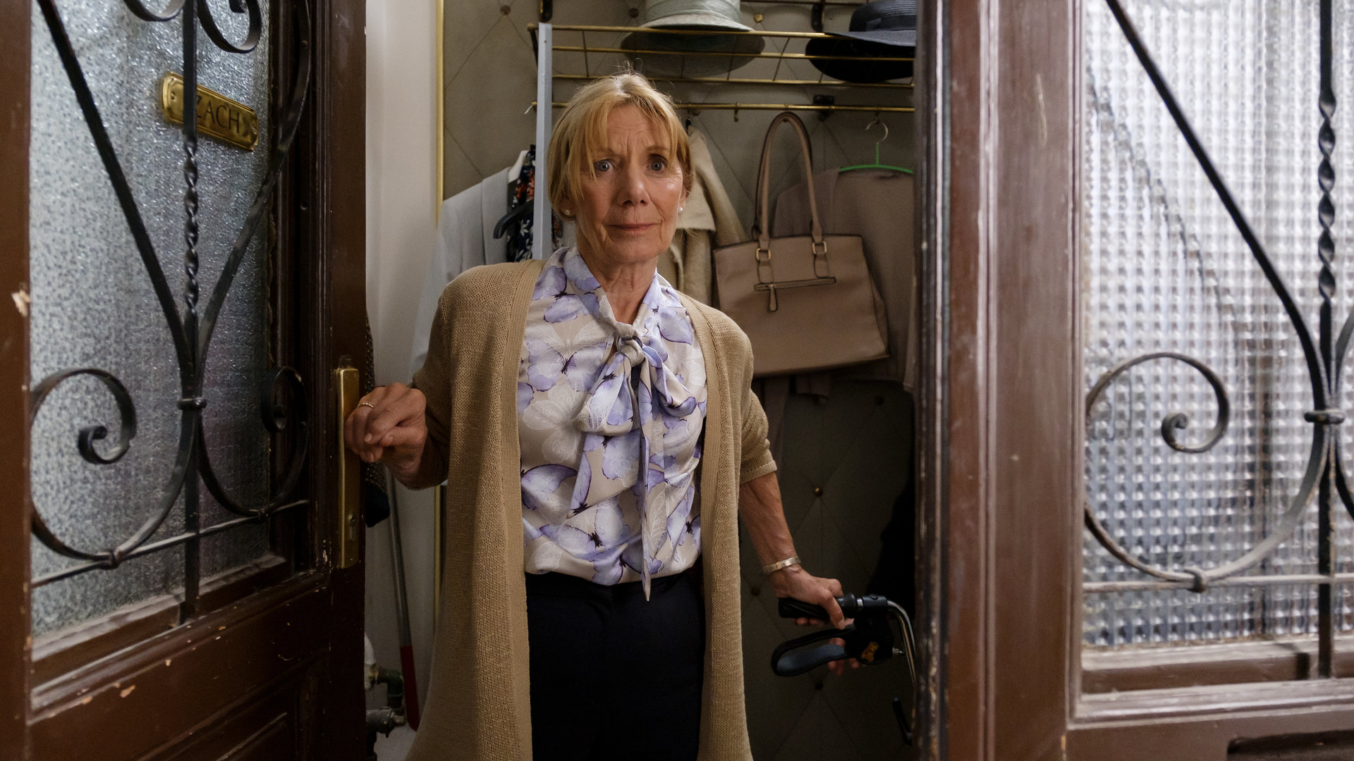 "SOKO Wien - Der gschupfte Ferdl": Wetty Zach (Erika Deutinger) steht in ihrer geöffneten Wohnungseingangstür. Mit ihrer rechten Hand hält sie sich an der Türklinke fest und mit der linken Hand an ihrem Rollator. Im Hintergrund ist ihre Wandgarderobe an der eine Handtasche hängt. Zwei Hüte liegen auf der Hutablage.