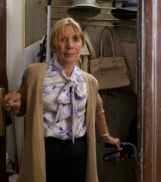 "SOKO Wien - Der gschupfte Ferdl": Wetty Zach (Erika Deutinger) steht in ihrer geöffneten Wohnungseingangstür. Mit ihrer rechten Hand hält sie sich an der Türklinke fest und mit der linken Hand an ihrem Rollator. Im Hintergrund ist ihre Wandgarderobe an der eine Handtasche hängt. Zwei Hüte liegen auf der Hutablage.