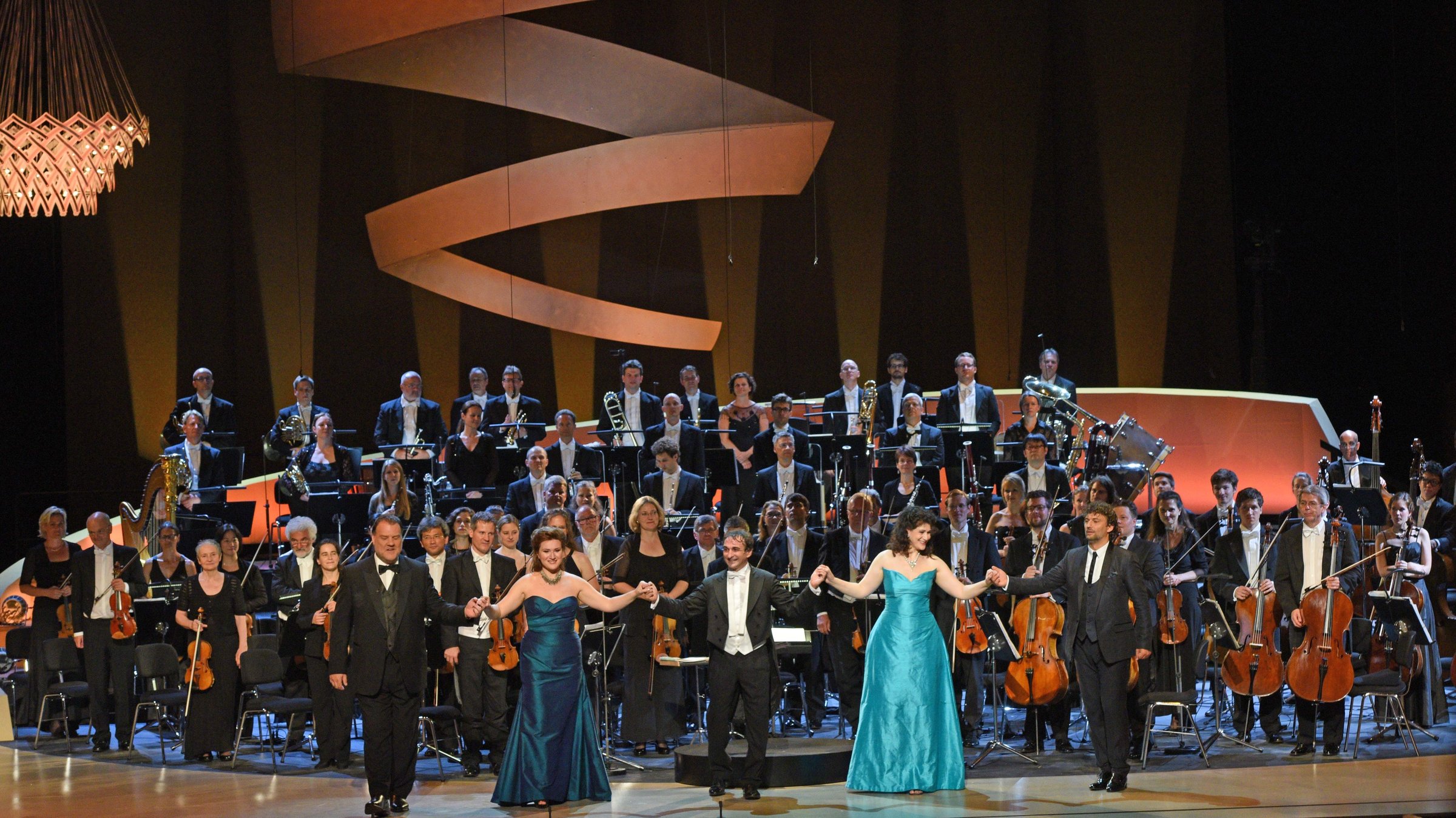 "Sommernachtsmusik (Sendetitel in 3sat: Gala aus dem Festspielhaus Baden-Baden)": Gruppenbild von Bryn Terfel (Bassbariton), Ekaterina Gubanova (Mezzosopran), Marco Armiliato (Dirigent), Anja Harteros (Sopran), Jonas Kaufmann (Tenor) vor der Badischen Staatskapelle auf der Bühne.