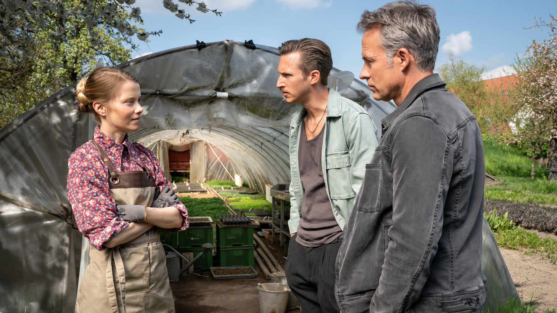 "SOKO Leipzig - Aus deutschen Landen": Mathilde Karnitz (Henrike von Kuick) steht mit verschränkten Armen Moritz Brenner (Johannes Hendrik Langer) und Jan Maybach (Marco Girnth) vor einem Foliengewächshaus gegenüber. Sie trägt Handschuhe und eine Schürze.