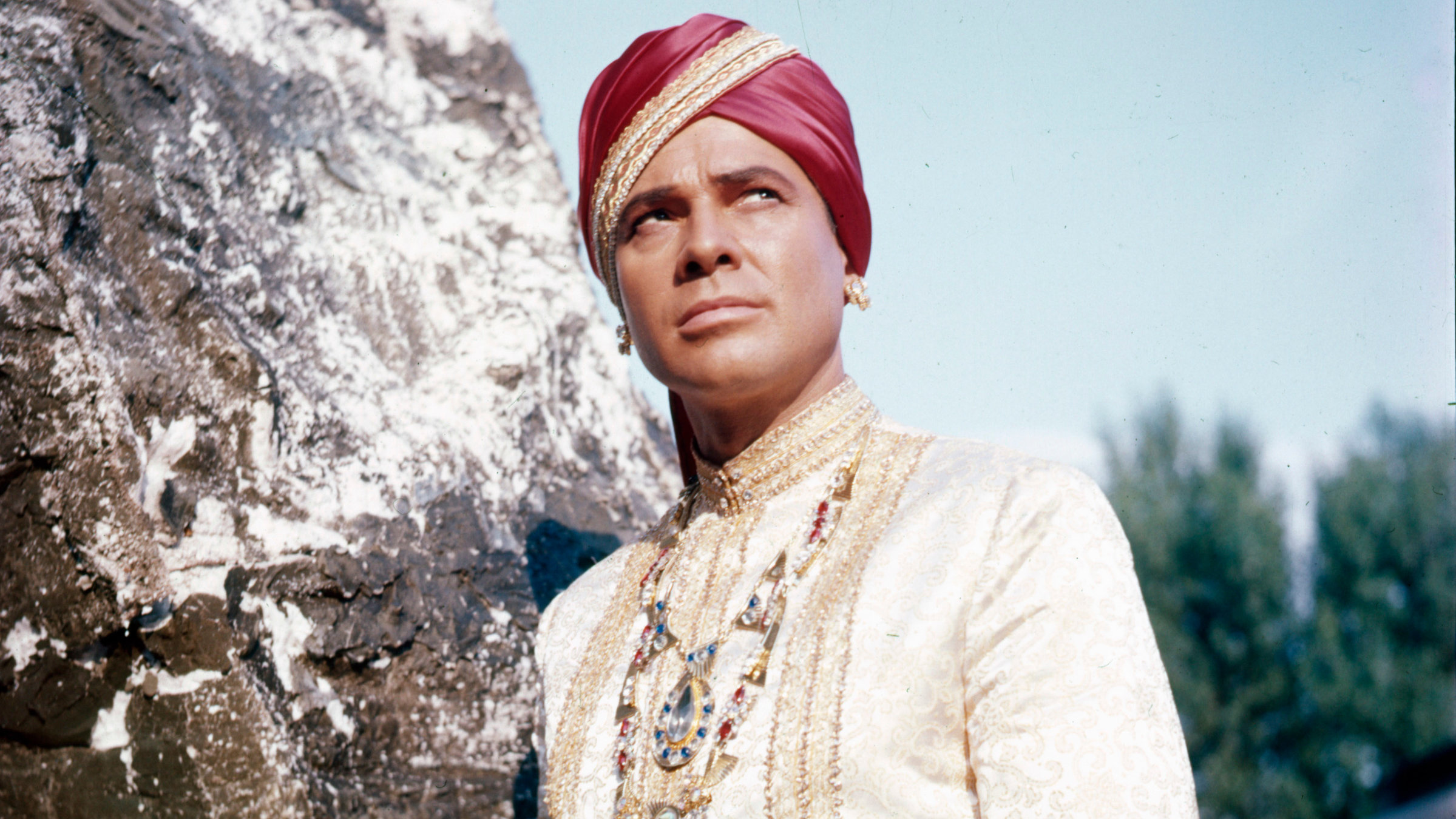 "Der Tiger von Eschnapur": Der Maharadscha von Eschnapur (Walther Reyer) trägt eine herrschaftliche weiße Tracht mit einer prunkvollen Halskette, einer roten Schärpe sowie einem roten Turban und lehnt wie in Pose stehend an einem Felsen. Sein skeptisch-versonnen wirkender Blick richtet sich nach schräg oben.