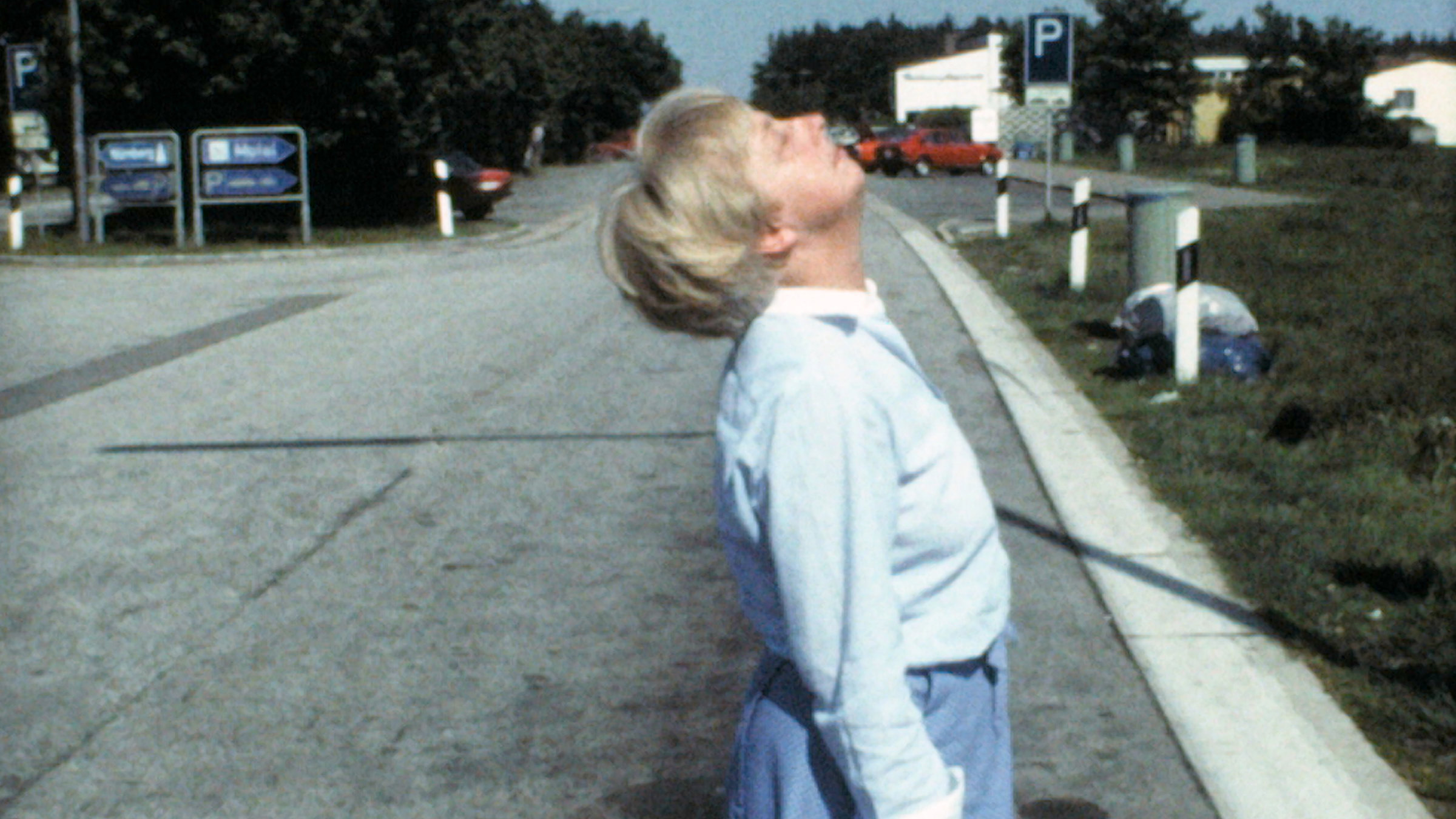 "Lydia":  Halbnahe Aufnahme einer Frau von der Seite, die auf dem Parkplatz einer Autobahn-Raststätte steht und sich mit geschlossenen Augen nach hinten beugt.