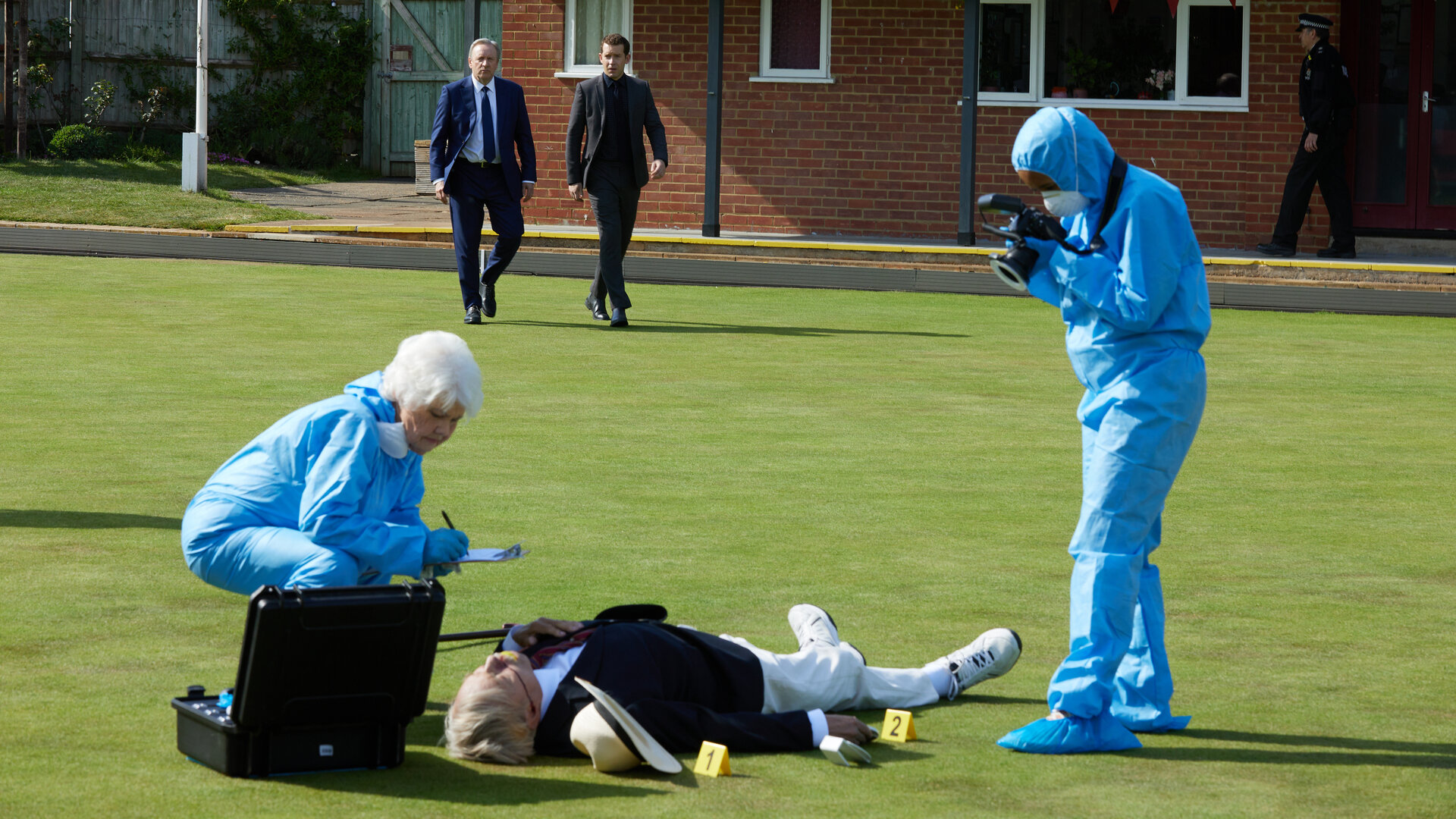 "Inspector Barnaby - Der Tod rollt auf dem Bowling Green": Wilf Worrell (Frazer Hines) liegt tot auf einer Rasenfläche. Fleur Perkins (Annette Badland), gekleidet in einem blauen Schutzanzug, kniet neben ihm, um ihn zu untersuchen. Ein weiterer Beamter (Komparse) in Schutzanzug steht rechts daneben und fotografiert den Fundort. Im Hintergrund nähern sich John Barnaby (Neil Dudgeon) und Jamie Winter (Nick Hendrix).