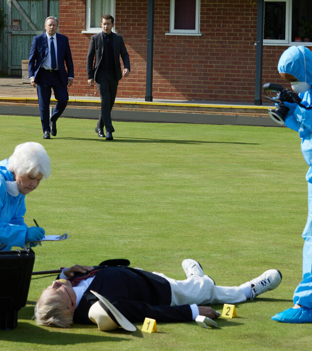 "Inspector Barnaby - Der Tod rollt auf dem Bowling Green": Wilf Worrell (Frazer Hines) liegt tot auf einer Rasenfläche. Fleur Perkins (Annette Badland), gekleidet in einem blauen Schutzanzug, kniet neben ihm, um ihn zu untersuchen. Ein weiterer Beamter (Komparse) in Schutzanzug steht rechts daneben und fotografiert den Fundort. Im Hintergrund nähern sich John Barnaby (Neil Dudgeon) und Jamie Winter (Nick Hendrix).