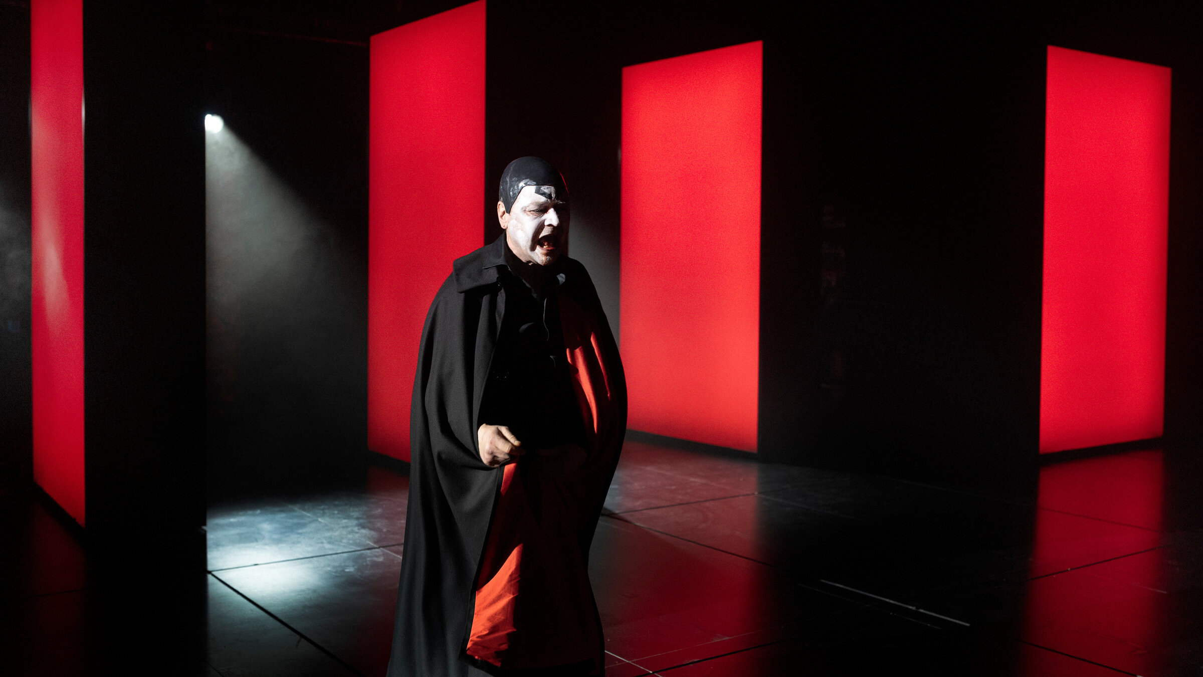 "Mephisto": Ein Mann im langen, dunklen Umhang, schwarzer kappe und weiß geschminktem Gesicht steht vor mehreren hohen, rechteckigen Paneelen, die in kräftigem Rot leuchten. Der restliche Bühnenraum ist dunkel.