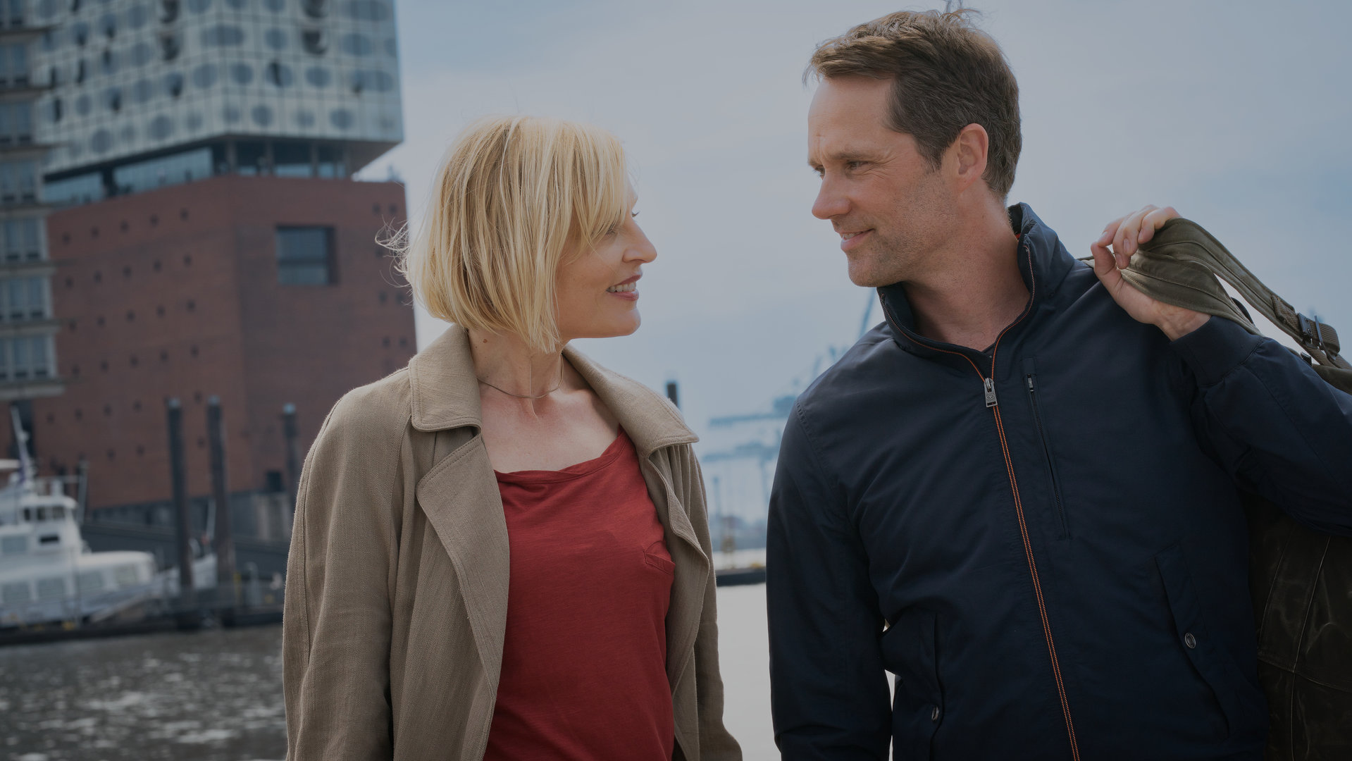 "Notruf Hafenkante - Gegenwind": Großaufnahme von Melanie Hansen (Sanna Englund) und Mattes Seeler (Matthias Schloo). Sie stehen nebeneinander und grinsen sich an. Beide sind in zivil. Im Hintergrund liegt die Hafenanlage mit Gebäuden.