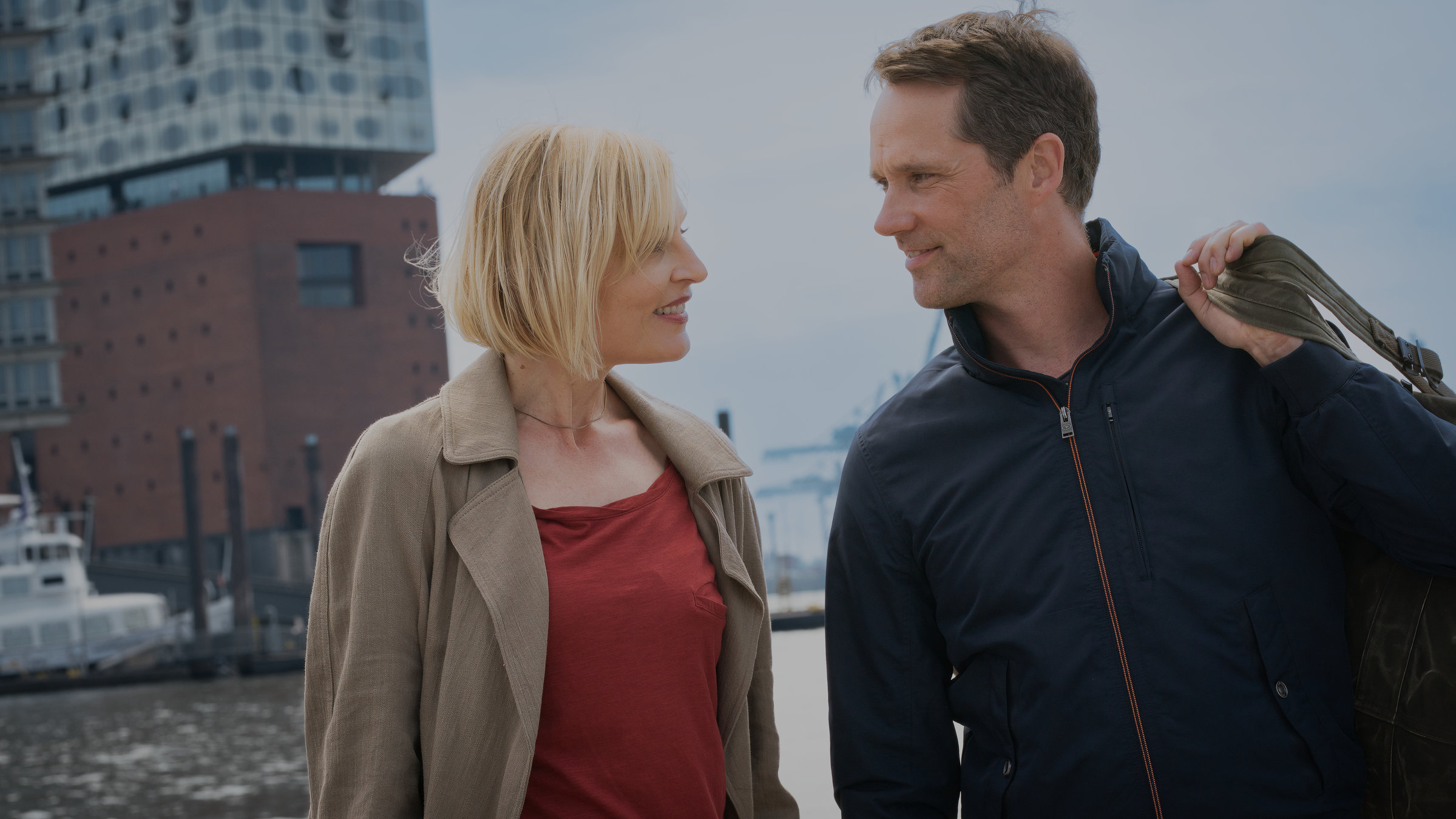 "Notruf Hafenkante - Gegenwind": Großaufnahme von Melanie Hansen (Sanna Englund) und Mattes Seeler (Matthias Schloo). Sie stehen nebeneinander und grinsen sich an. Beide sind in zivil. Im Hintergrund liegt die Hafenanlage mit Gebäuden.