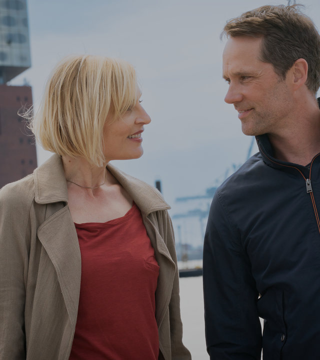 "Notruf Hafenkante - Gegenwind": Großaufnahme von Melanie Hansen (Sanna Englund) und Mattes Seeler (Matthias Schloo). Sie stehen nebeneinander und grinsen sich an. Beide sind in zivil. Im Hintergrund liegt die Hafenanlage mit Gebäuden.