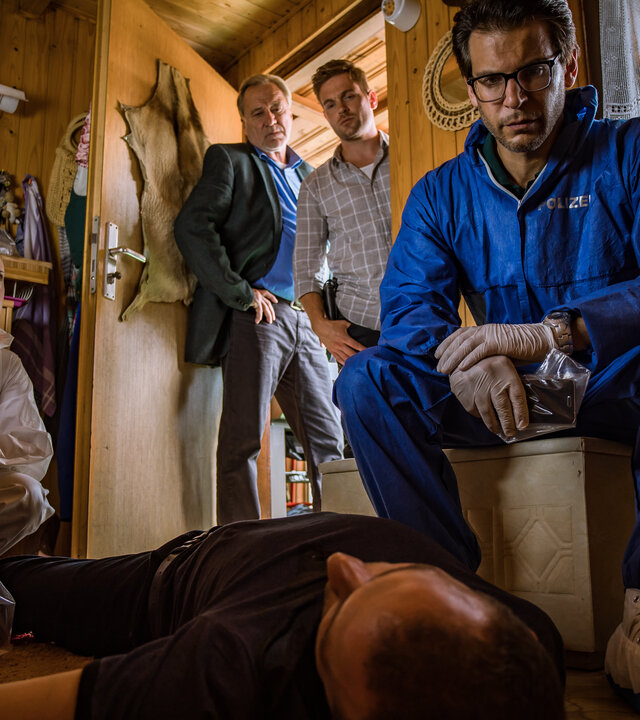 "SOKO München - Wer anderen eine Grube gräbt": Das Team der SOKO ist an einem Tatort in einer kleinen Gartenhütte. Ein toter Mann liegt in der Mitte des Raumes auf dem Rücken. Neben ihm steht ein Gewehr, das in einer Plastiktüte steckt und der Koffer des Spurensicherung. Rechts von ihm sitzt Dr. Weissenböck (Florian Odendahl) auf einer weißen Truhe und sieht ihn sich nachdenklich an. Dr. Weissenböck trägt einen blauen Schutzanzug, sowie weiße Gummihandschuhe und weiß-blaue Schutzüberzüge für die Schuhe. Er hält einen Plastikbeutel in den Händen, in dem sich ein schwarzes Handy befindet. Am Fußende des Mannes sitzt Billie Curio (Sina Reiß) und blickt zu Dr. Weissenböck. Billie Curio trägt einen weiß-orangenen Schutzanzug und lila Gummihandschuhe. Dominik Morgenstern (Joscha Kiefer) und Arthur Bauer (Gerd Silberbauer) stehen in der Tür der Gartenhütte und betrachten die Leiche des Mannes.