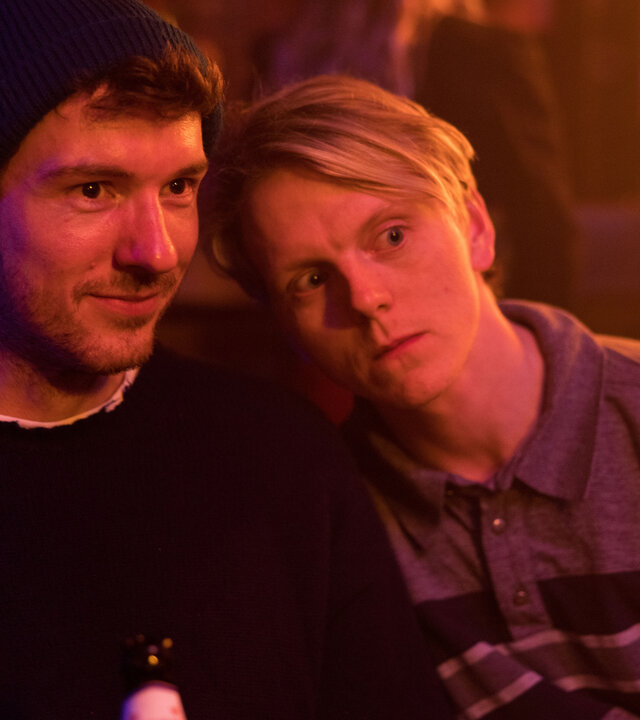 "This Is Gonna Be Great - Freunde": David (Rein Mulder) und Rob (Jonas Strand Gravli) stehen nebeneinander in einem bunt ausgeleuchteten Raum. David lächelt, Rob neigt den Kopf in seine Richtung.