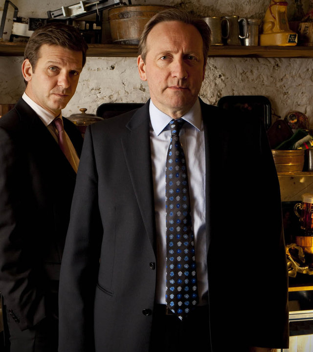 "Inspector Barnaby - Ein Funke genügt": Die beiden schick gekleideten Ermittler stehen in einem kellerähnlichen Raum, der nur grob verputzt ist. An der Wand und neben Barnaby stehen offene Holzregale, auf denen allerlei Krempel steht. Beide Männer blicken in Richtung Kamera.