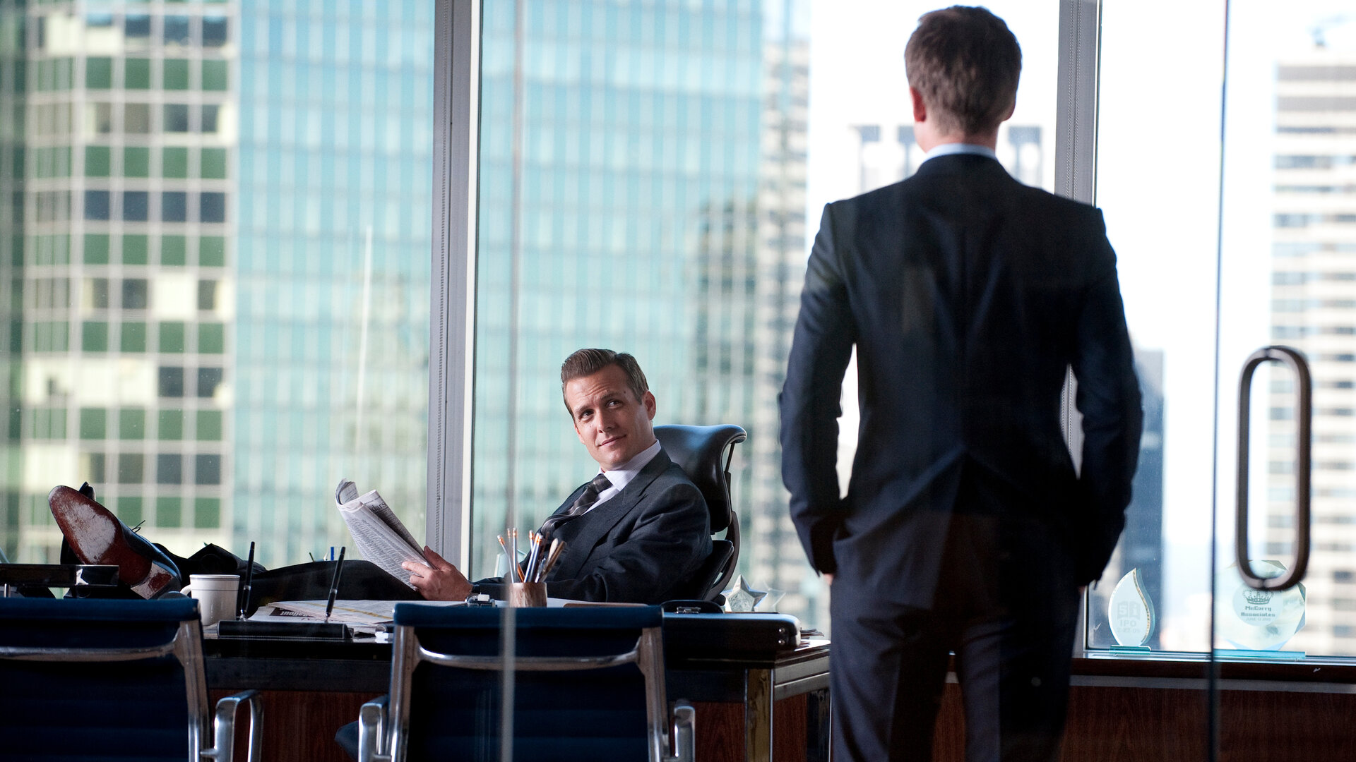 "Suits - Doppeltes Spiel": Harvey Specter (Gabriel Macht) sitzt entspannt mit hochgelegten Füßen an seinem Schreibtisch und blickt Mike Ross (Patrick J. Adams) an, der ihm mit dem Rücken zur Kamera gegenübersteht. Im Hintergrund geben große Fenster den Blick auf die Skyline frei.