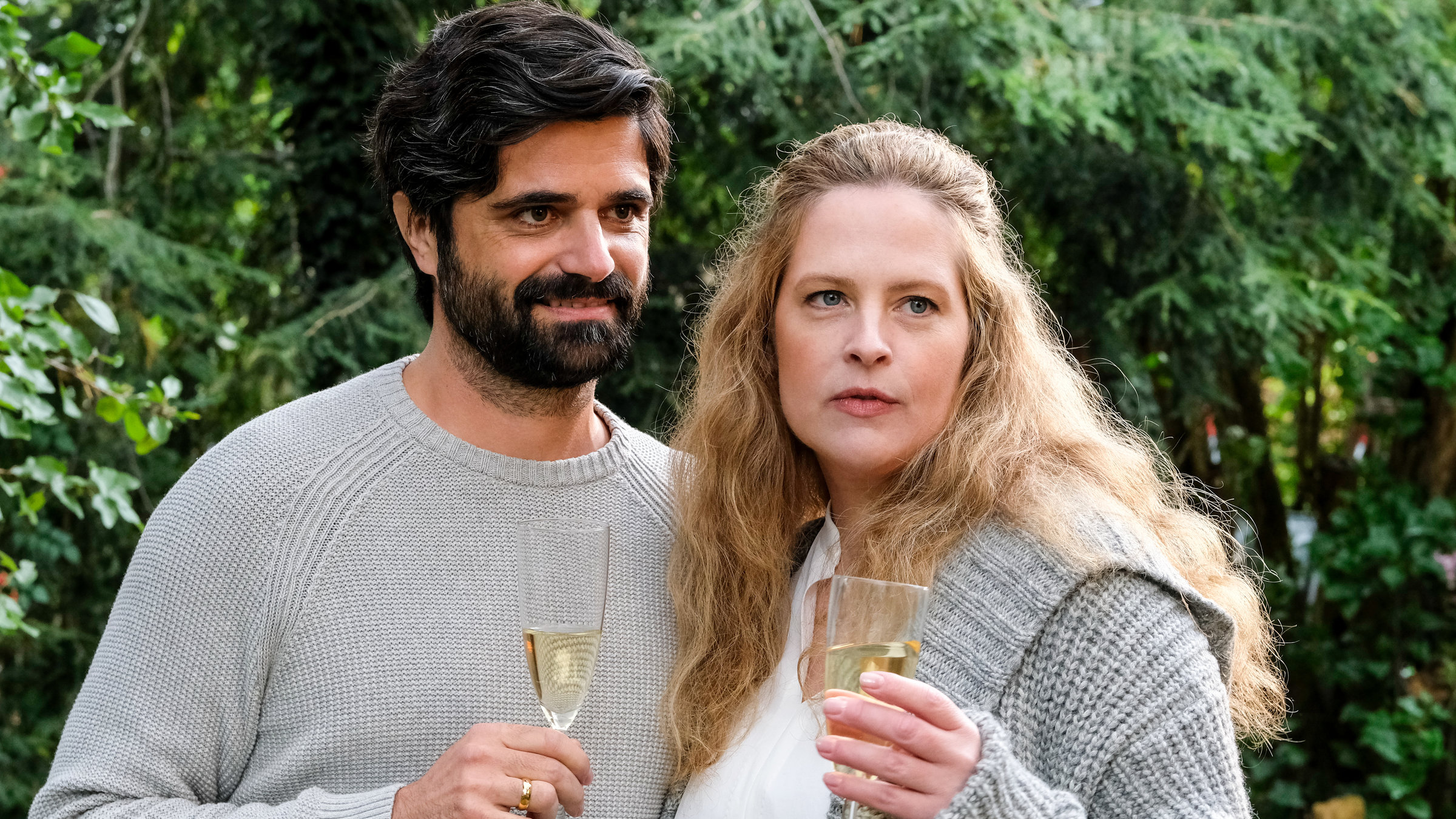 "Zum Glück zurück": Anne (Diana Amft) und Nils (Maximilian Grill) stehen Arm in Arm. Beide haben ein gefülltes Sektglas in der Hand. Nils trägt einen Wollpullover, Anne eine Bluse mit einer warmen Strickjacke darüber. Im Hintergrund sind Büsche und Nadelbäume zu sehen.