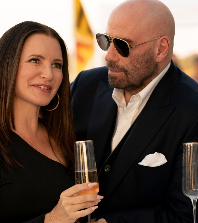 "Cash Out - Zahltag": Mason Goddard (John Travolta) und Amelia (Kristin Davis) stehen lächelnd in Abendgarderobe an einer Bar nah nebeneinander. Amelia hält ein halbvolles Sektglas in der Hand, vor Mason steht ein leeres. Im Hintergrund sind verschwommen weitere Gäste zu sehen.