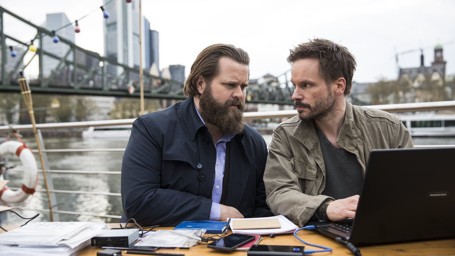 "Ein Fall für zwei - Das schwarze Schaf": Benni Hornberg (Antoine Monot, Jr.) und Leo Oswald (Wanja Mues) sitzen an Deck von Leos Hausboot. Leo hat einen Laptop vor sich und schaut Benni an. Im Hintergrund die Frankfurter Skyline.