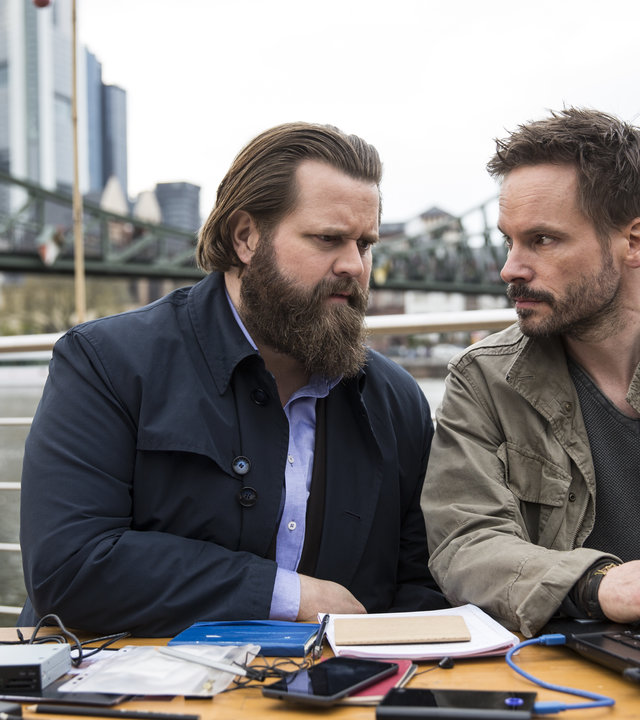 "Ein Fall für zwei - Das schwarze Schaf": Benni Hornberg (Antoine Monot, Jr.) und Leo Oswald (Wanja Mues) sitzen an Deck von Leos Hausboot. Leo hat einen Laptop vor sich und schaut Benni an. Im Hintergrund die Frankfurter Skyline.